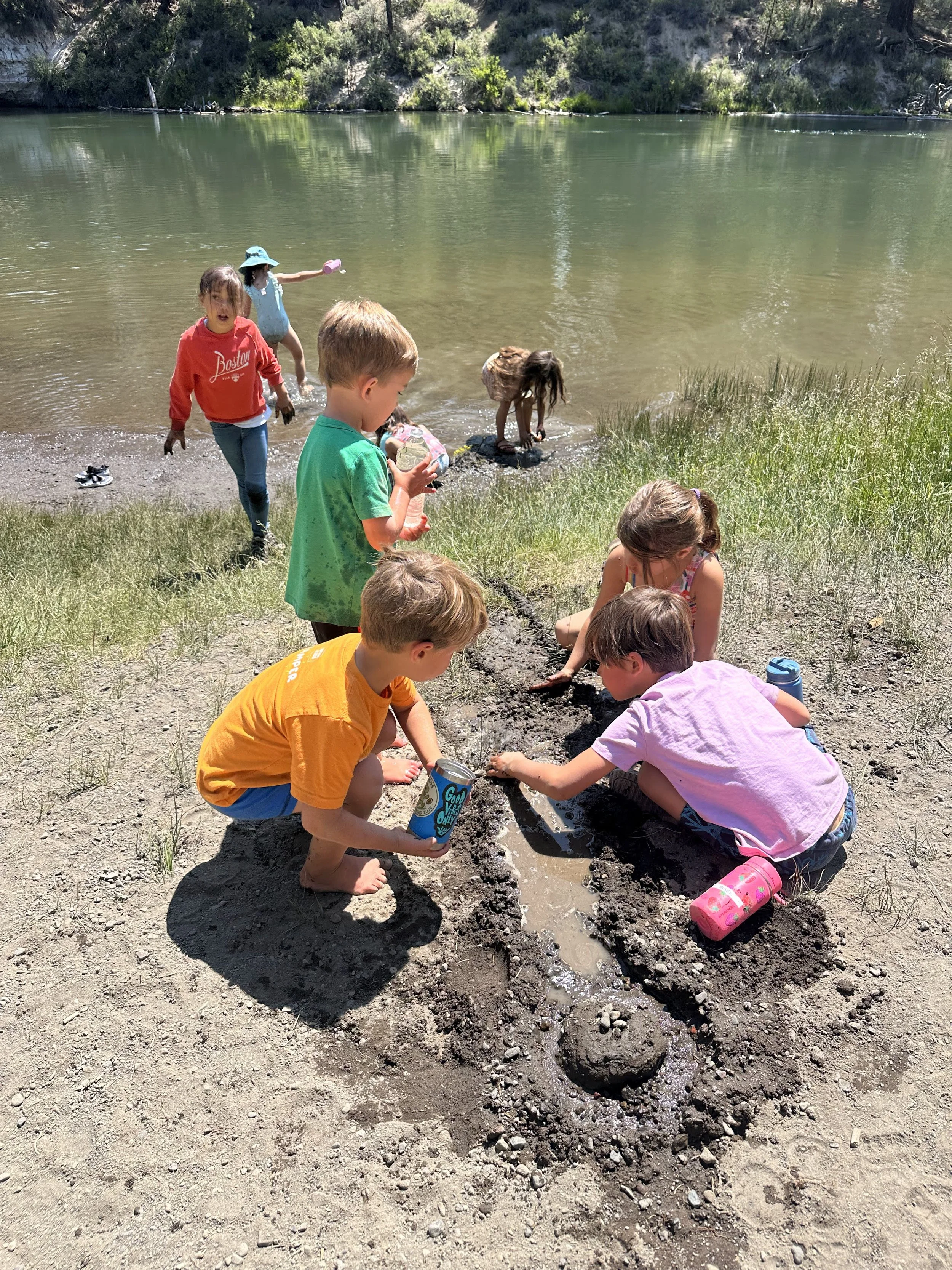 Children playing and digging in the dirt near a lake, with some splashing in the water and others building a mud structure.