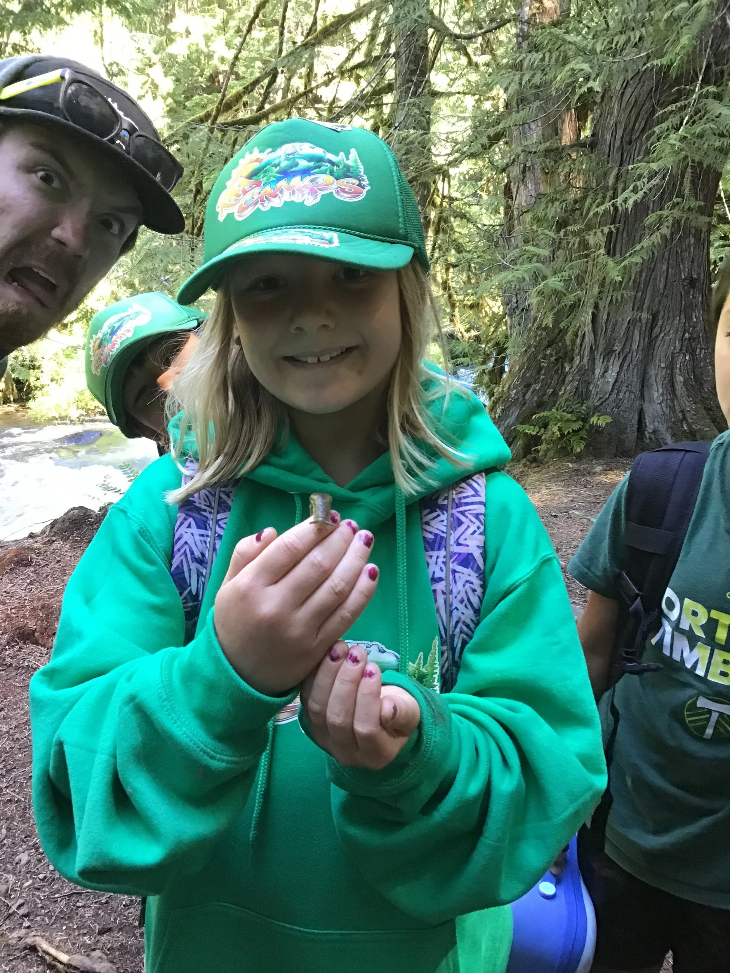 Child holding a small turtle in a forested area with other children and an adult nearby.