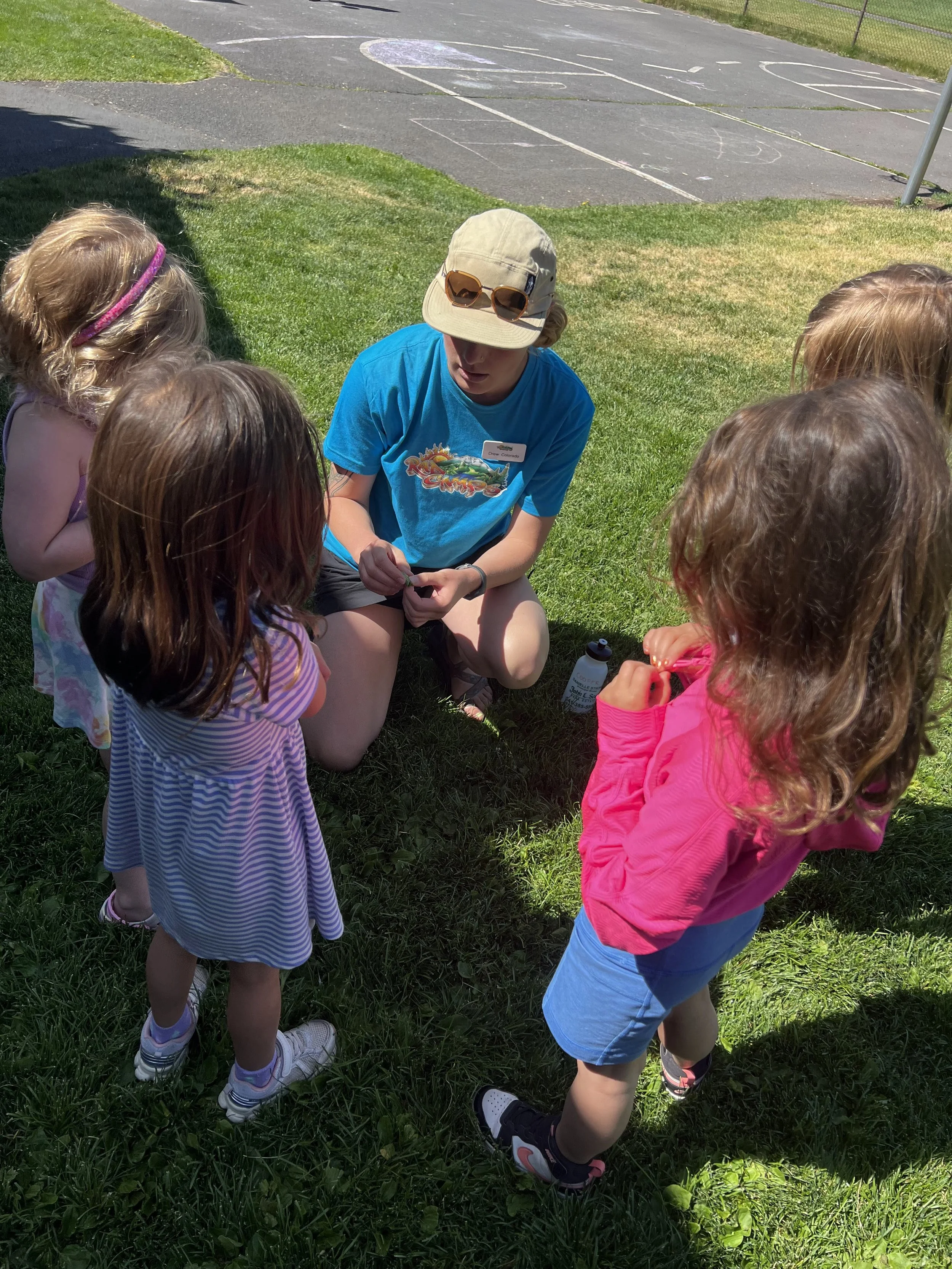 A female camp counselor kneeling on the grass and talking to five young girls gathered around her outdoors. The girls are listening attentively.