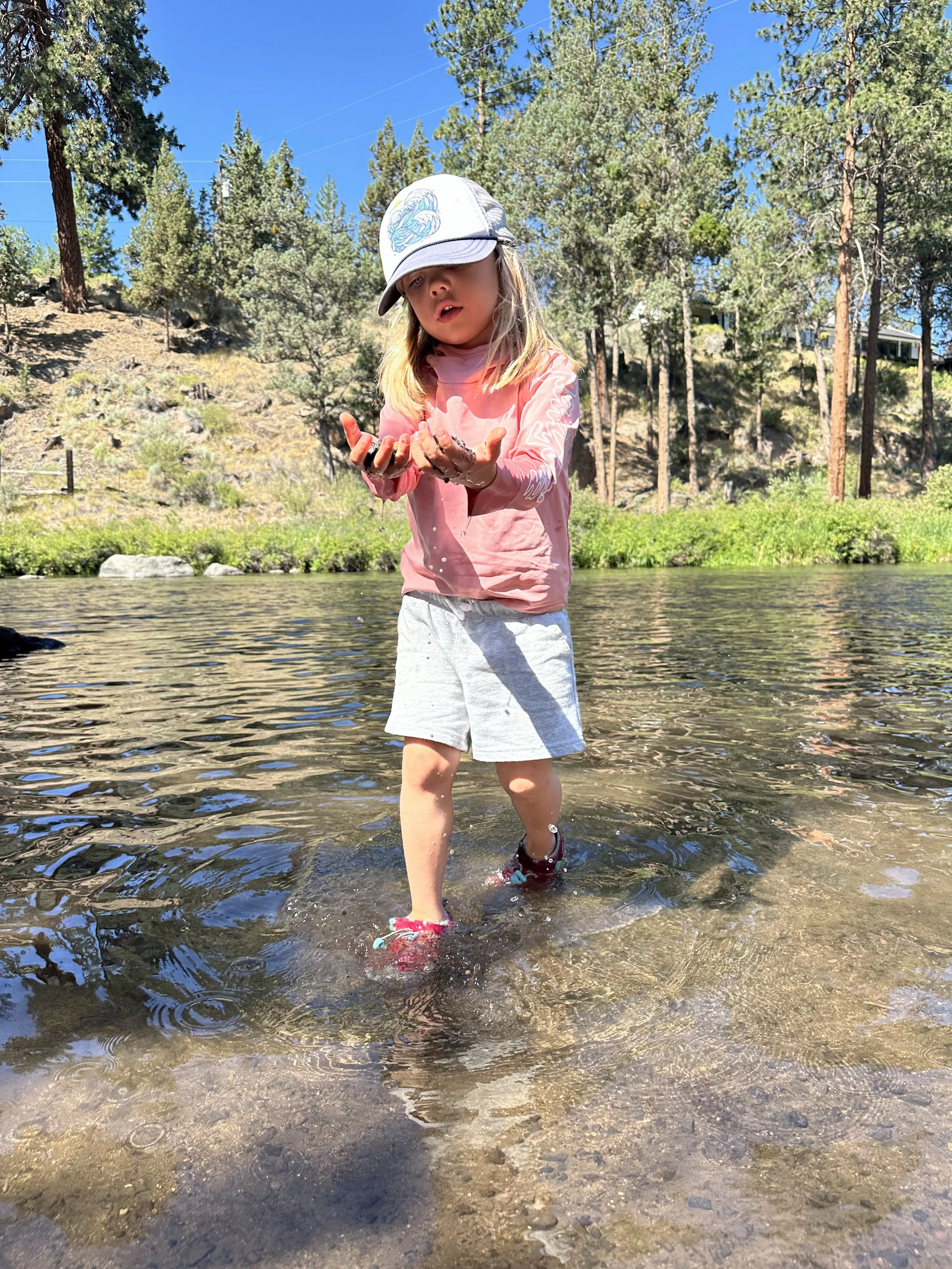 A young girl walking through a shallow river, wearing a pink long-sleeve shirt, gray shorts, a white cap with a blue design, and red water shoes, surrounded by trees and a clear blue sky.