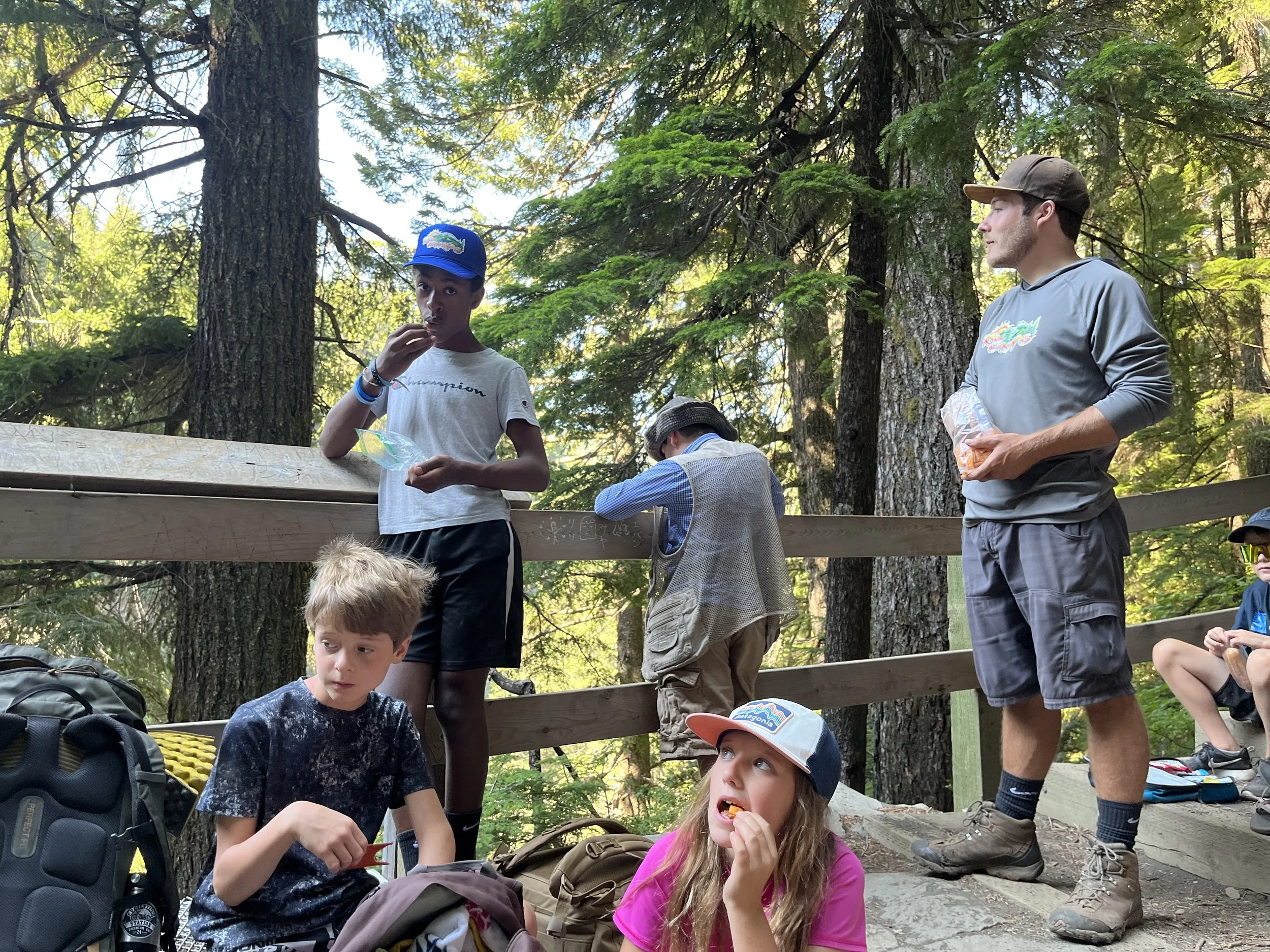 Group of children and a young adult sitting and standing on a wooden platform in a forest, some eating snacks and talking.