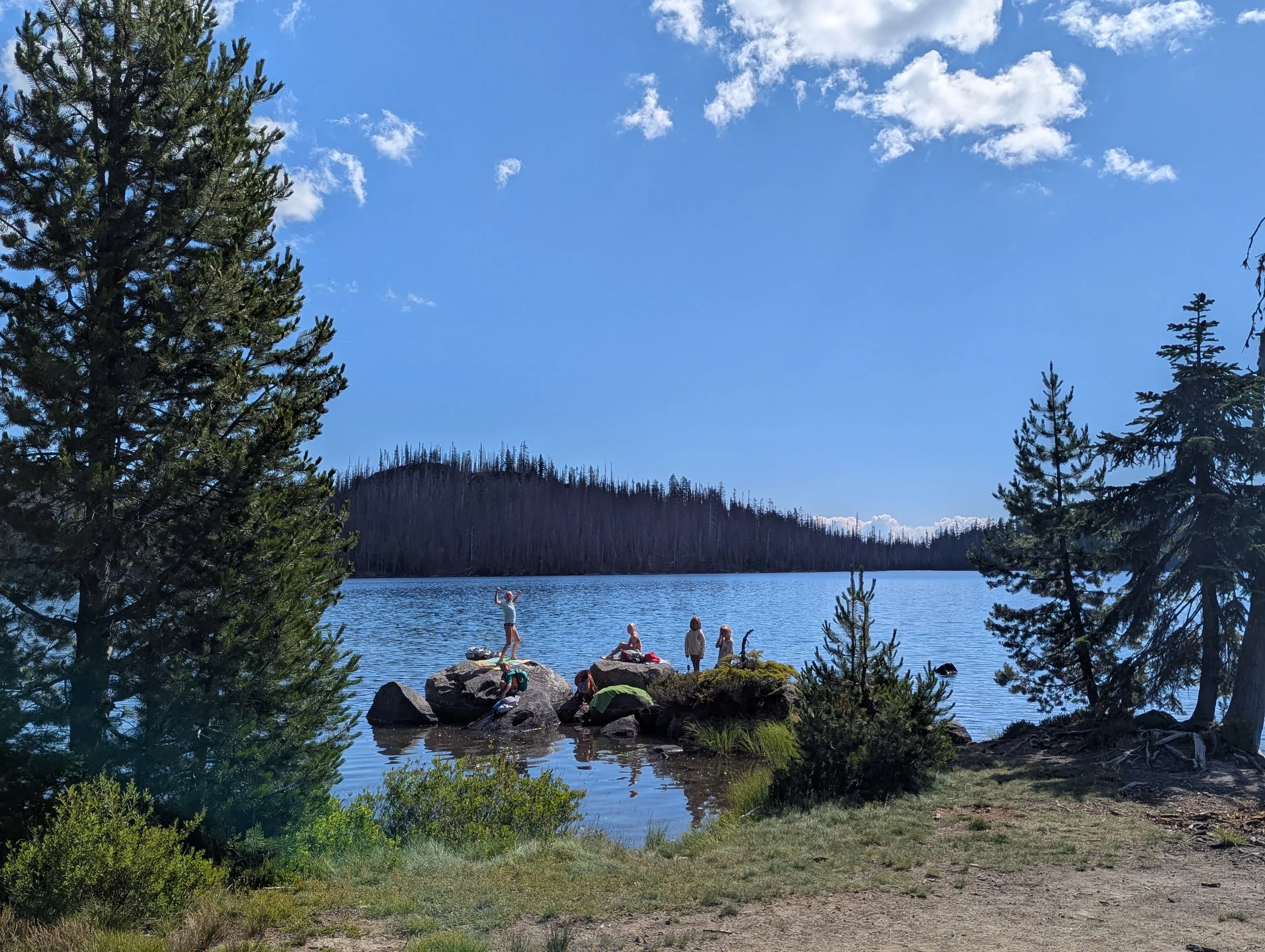 People relaxing on rocks by a lake under a clear sky with some scattered clouds, surrounded by pine trees and forested hills in the distance.