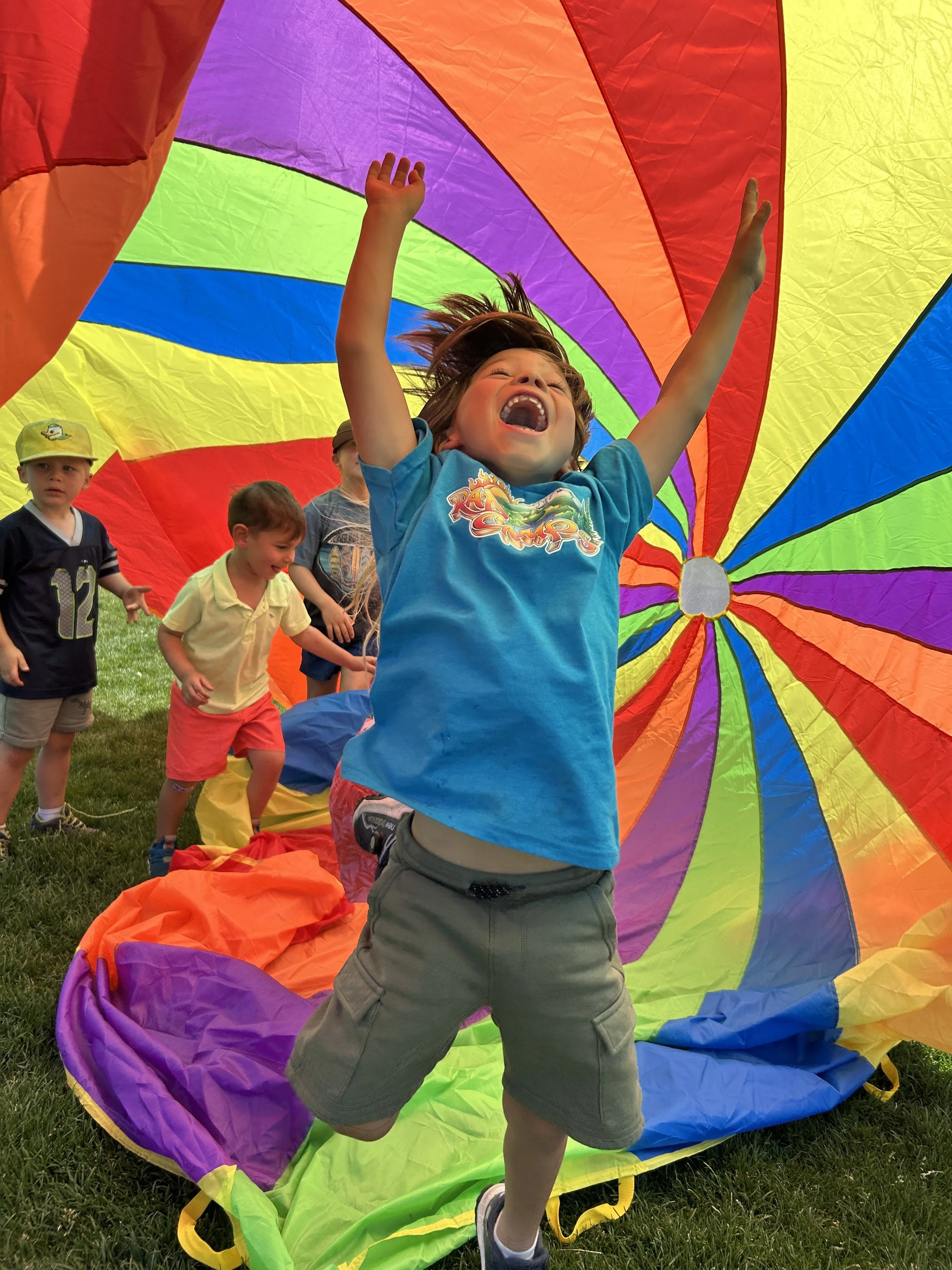 Children playing with a rainbow-colored parachute outdoors, with a boy in the foreground jumping with arms raised and the parachute draped behind him.