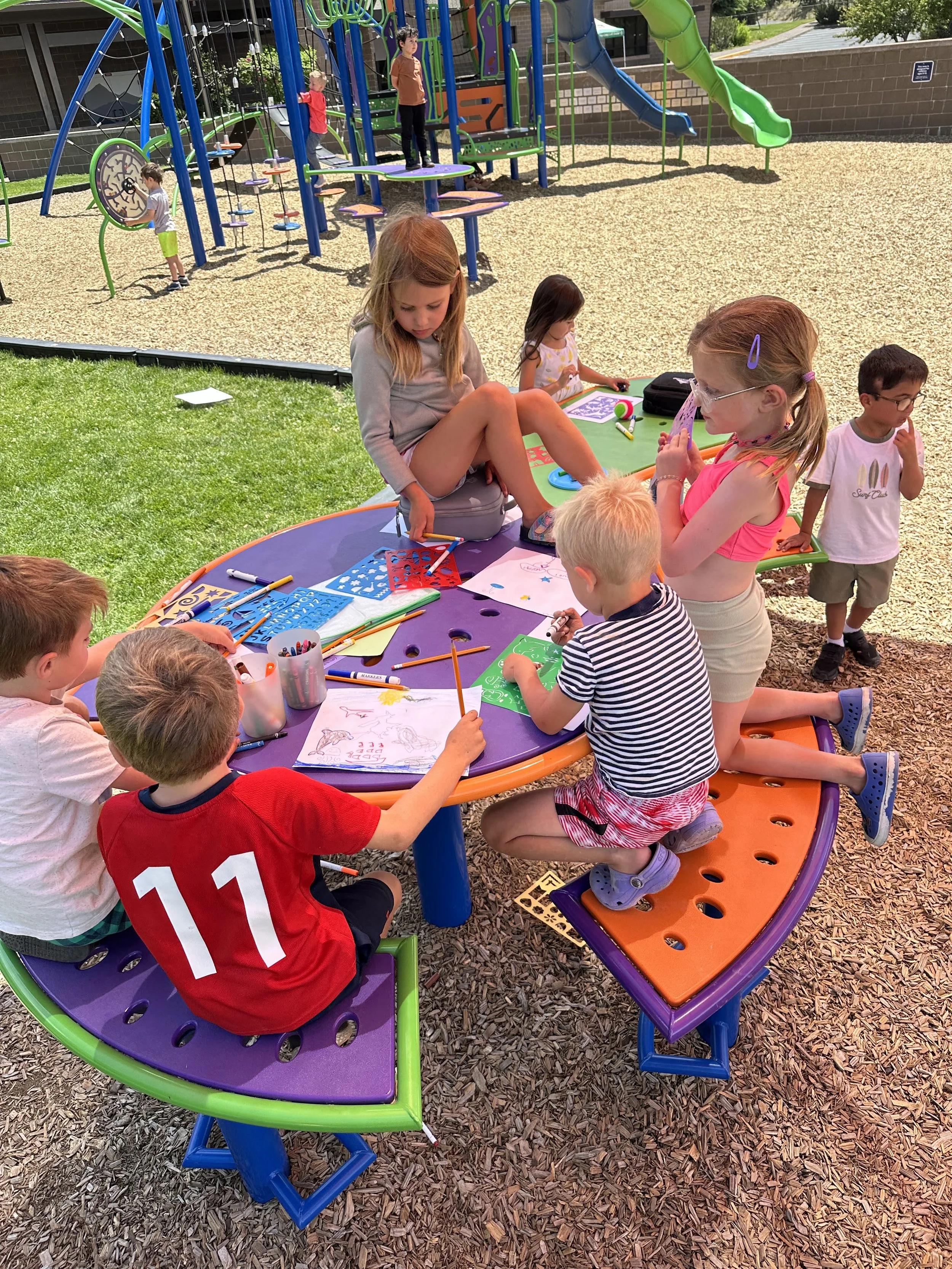 Children drawing and coloring at a round purple table outdoors with a playground in the background.