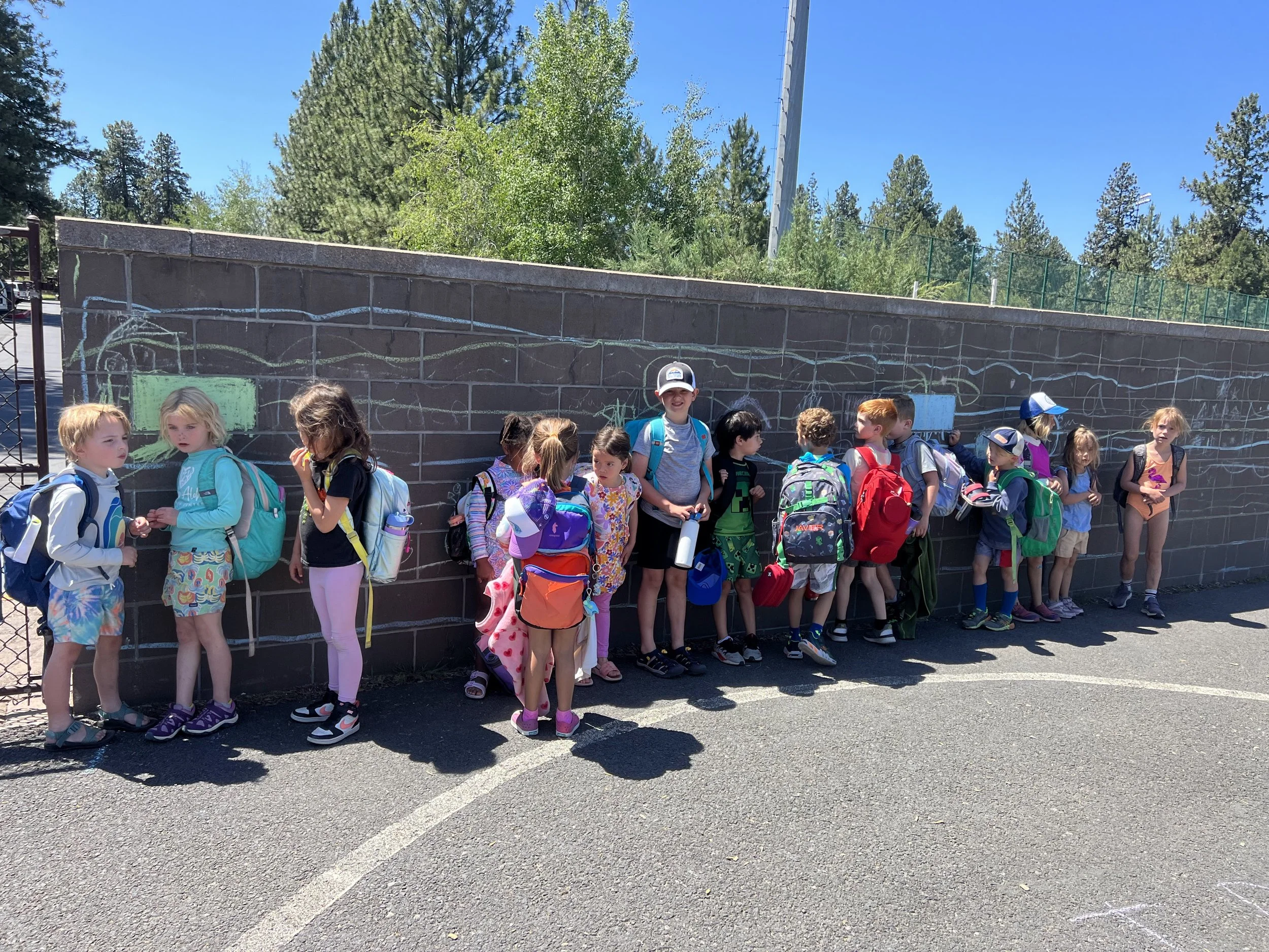A group of young children with backpacks standing against a black brick wall outdoors on a sunny day, pre-lineup.