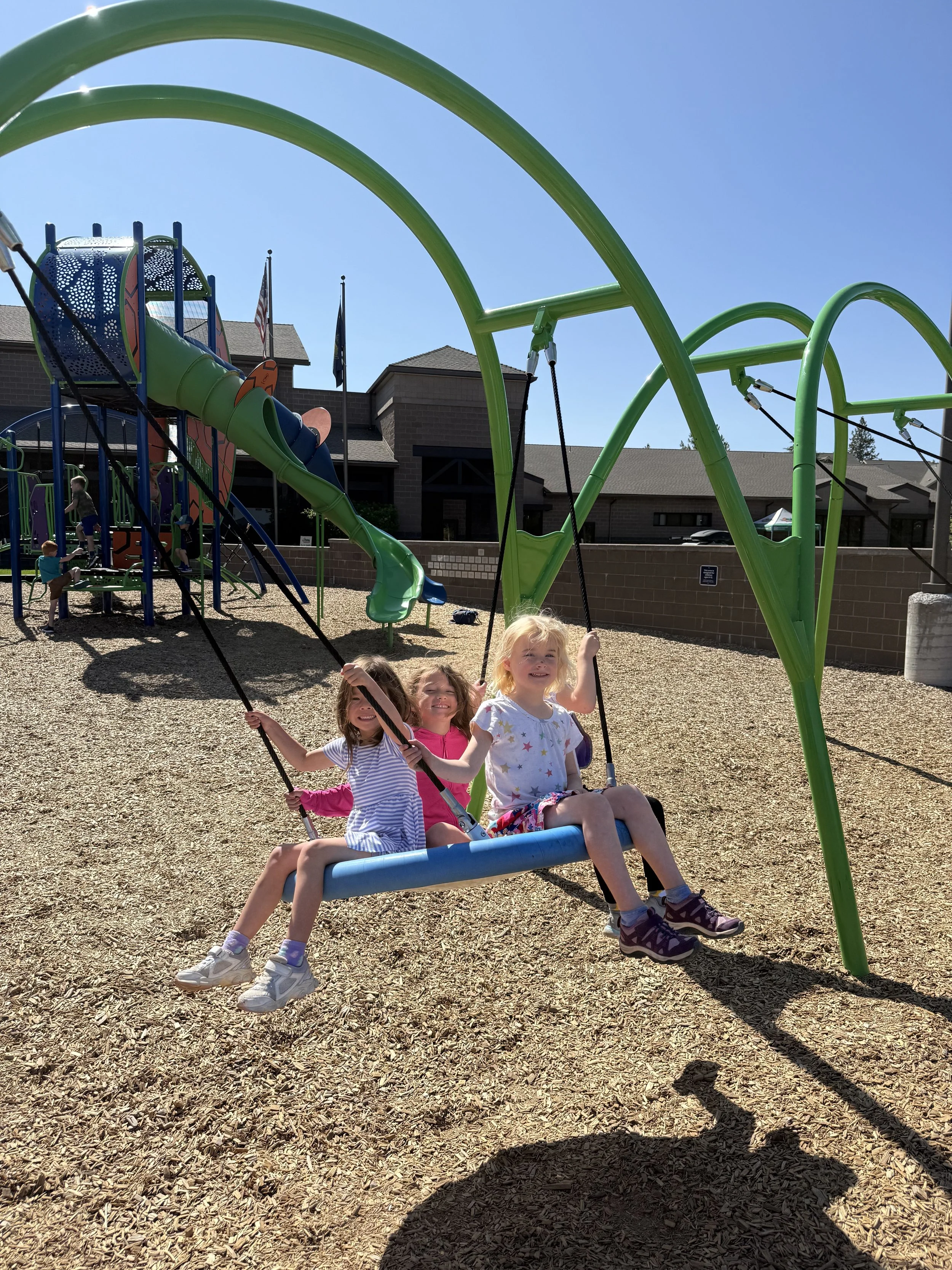 Three children playing on a swing at a playground with a green and blue slide in the background on a sunny day.