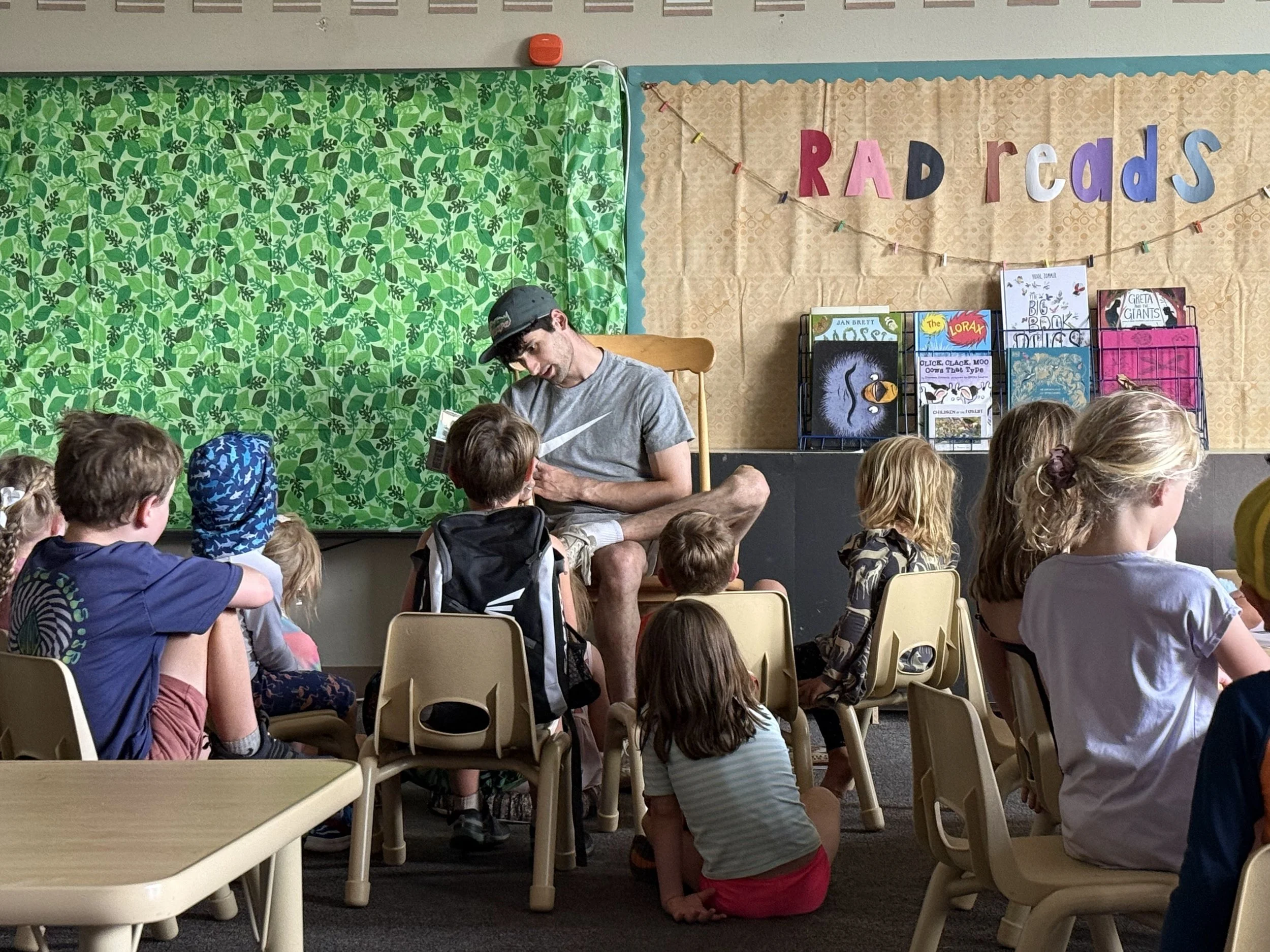 A young man reading a book to a group of children in a classroom. The children are sitting on small chairs and the young man is sitting on a larger wooden chair. The classroom has a green leafy curtain and a display with books on it. The background h