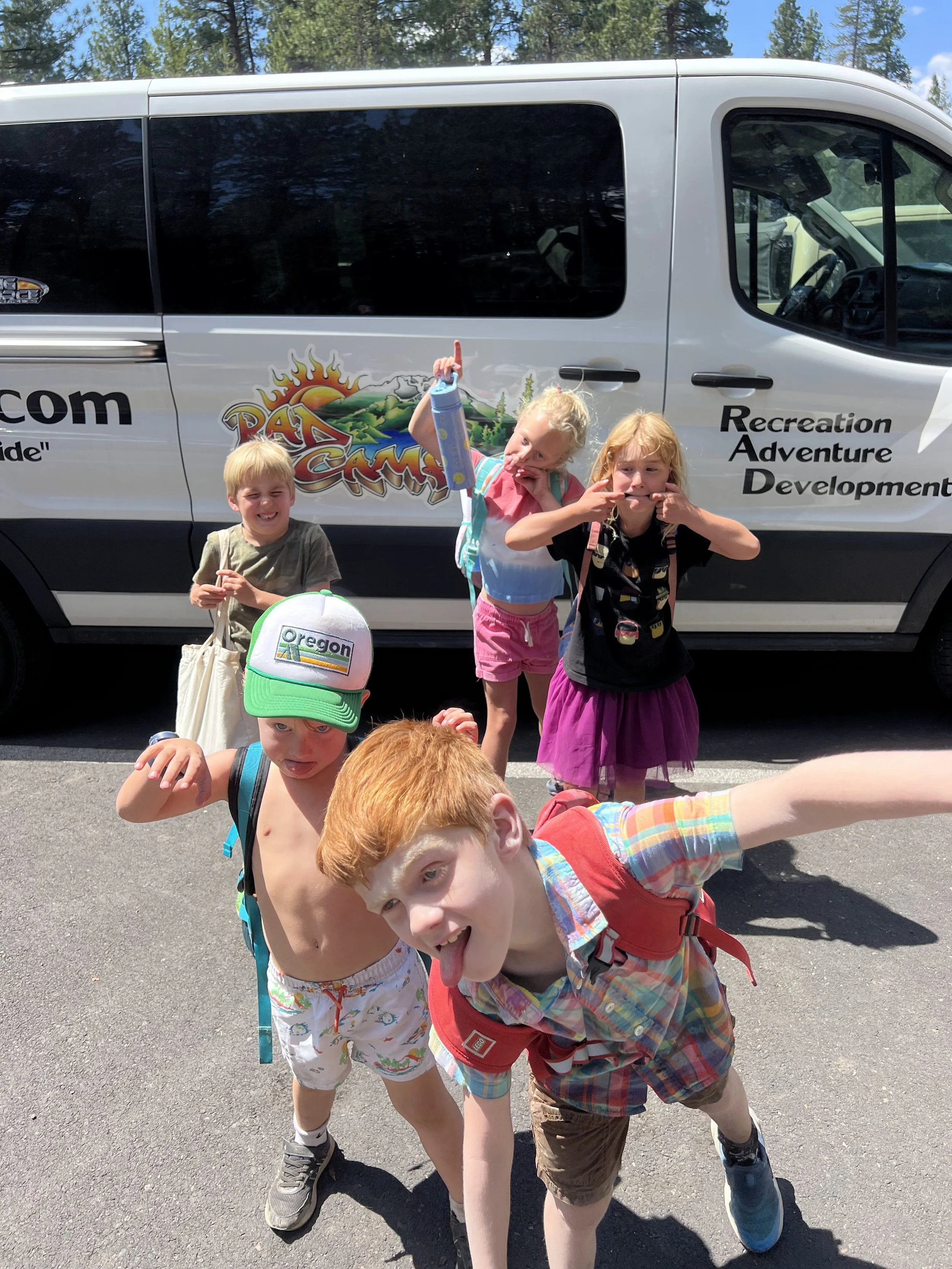 Five children with backpacks posing in front of a van with a colorful logo that says 'Recreation, Adventure, Development'.