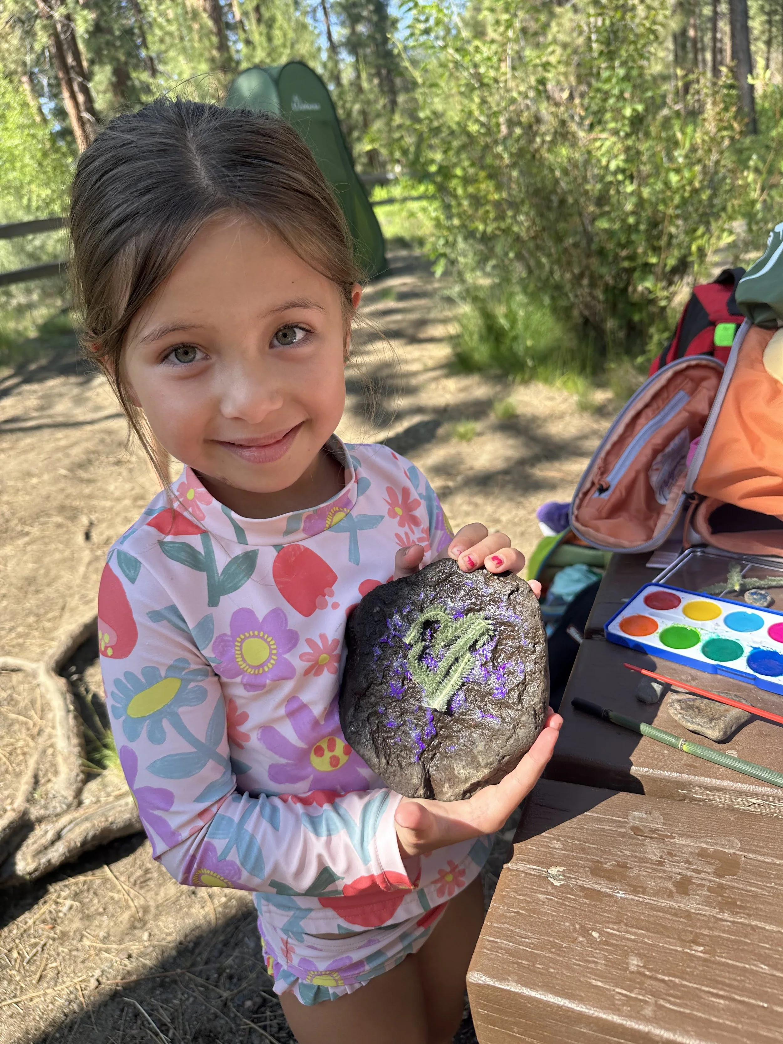 A young girl outdoors holding a volcanic rock decorated with colorful chalk drawings. She is smiling and standing near a table with watercolor paints and brushes, with trees and a dirt path in the background.