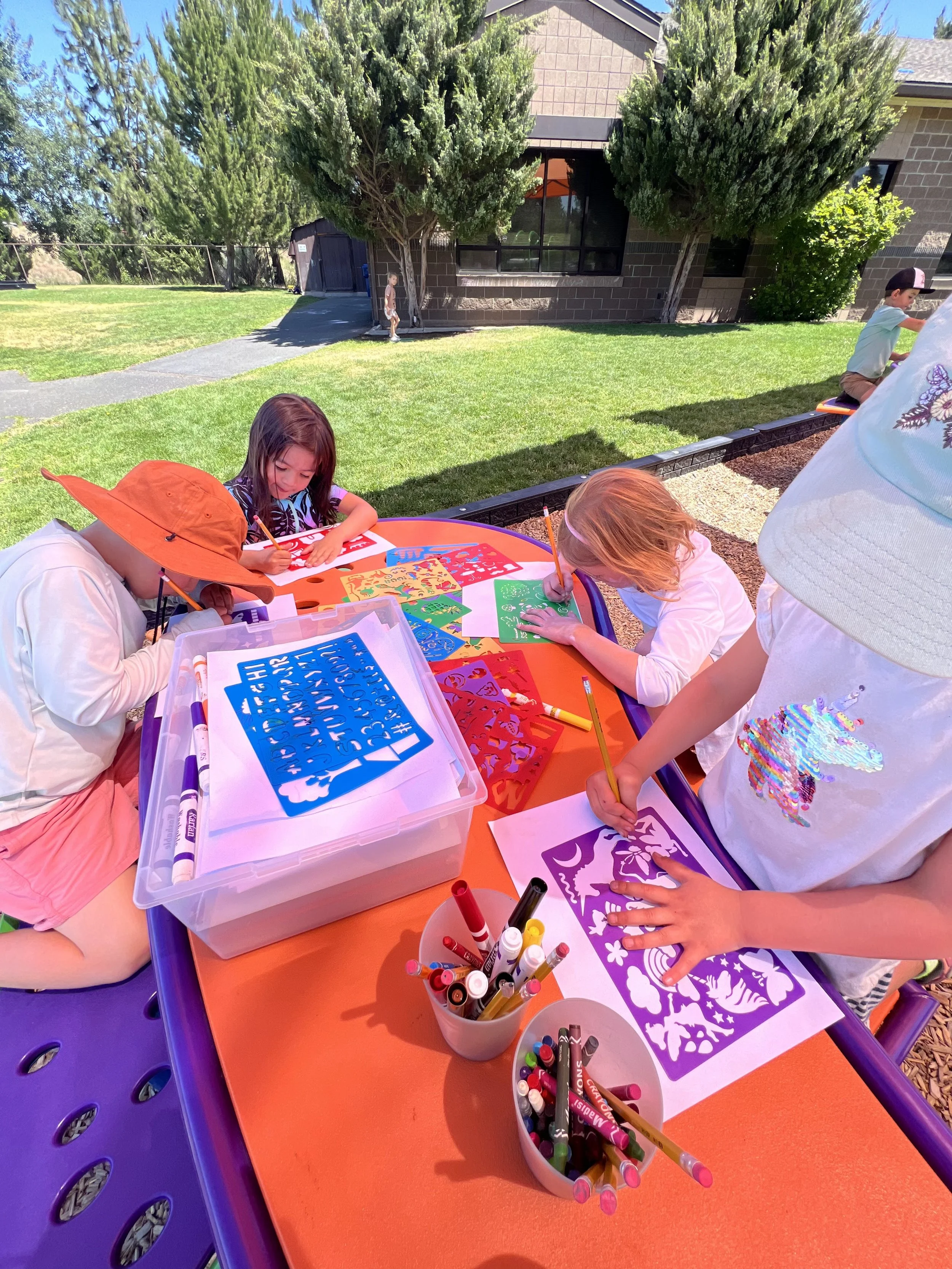 Children are sitting at an outdoor table engaged in coloring and arts and crafts activities with paper, markers, and stencils. The scene is in a backyard with green grass, trees, and a house in the background, on a sunny day.