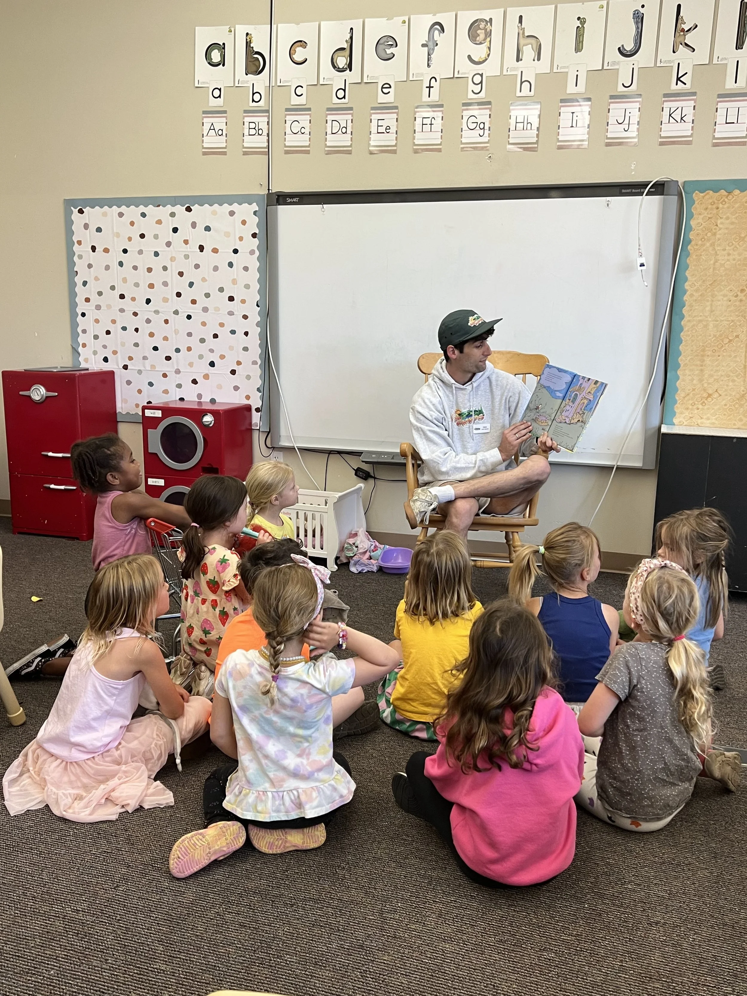 A man reading a children's book to a group of children seated on the floor in a classroom.