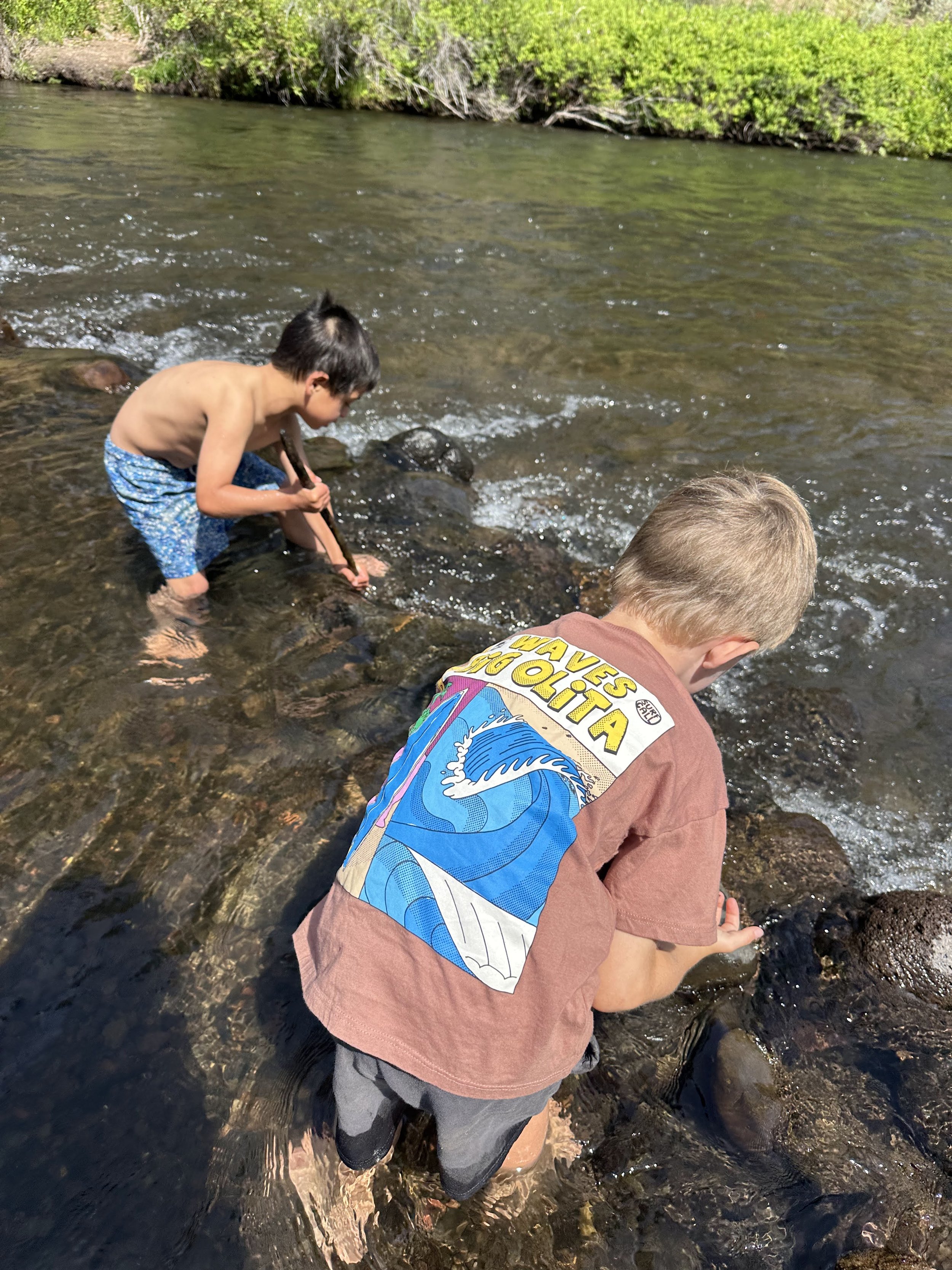 Two young boys are playing in a shallow river, kneeling on rocks and splashing water on a sunny day.
