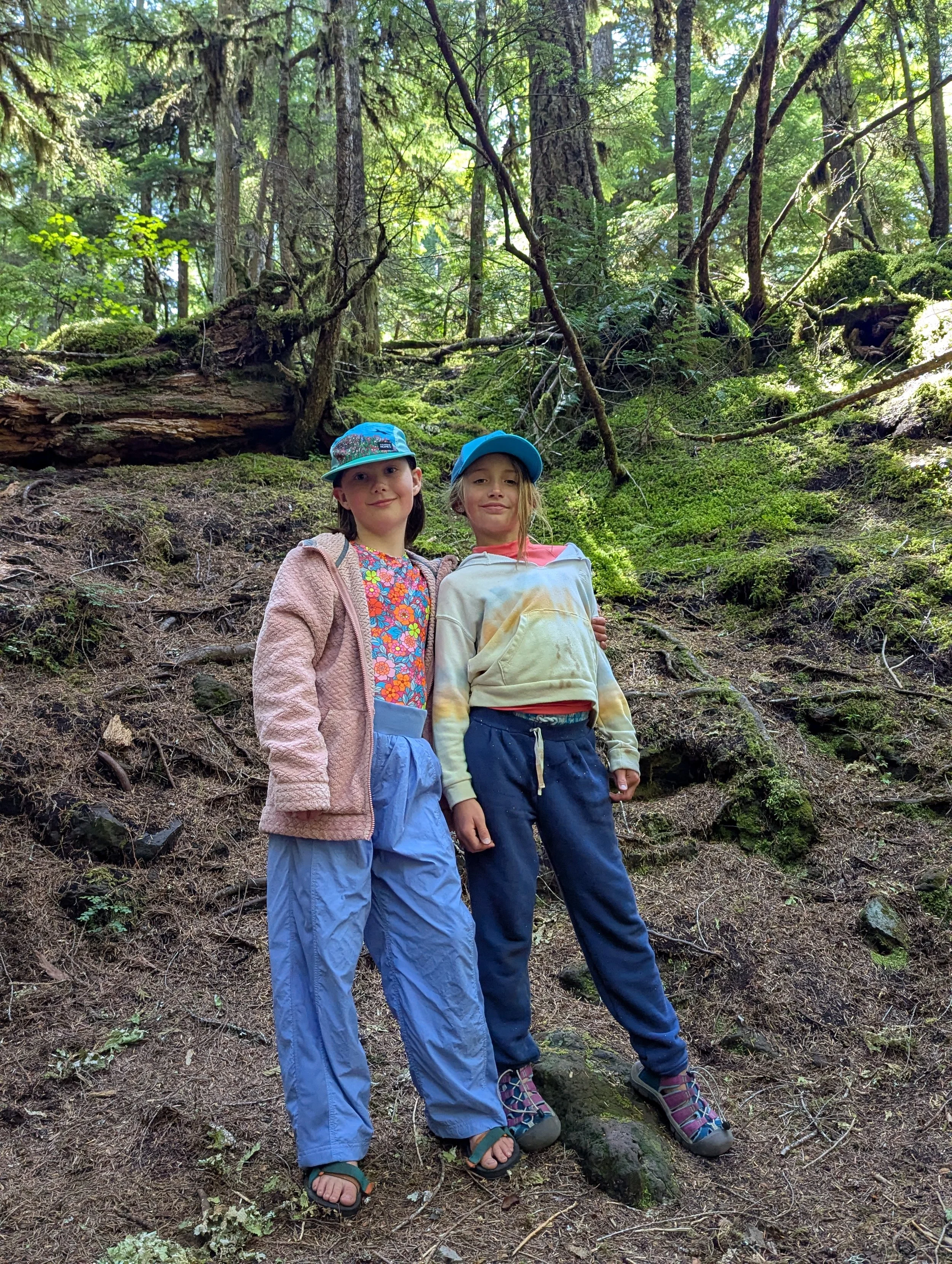 Two young girls stand together on a forest trail surrounded by trees and moss, wearing outdoor clothing and caps.