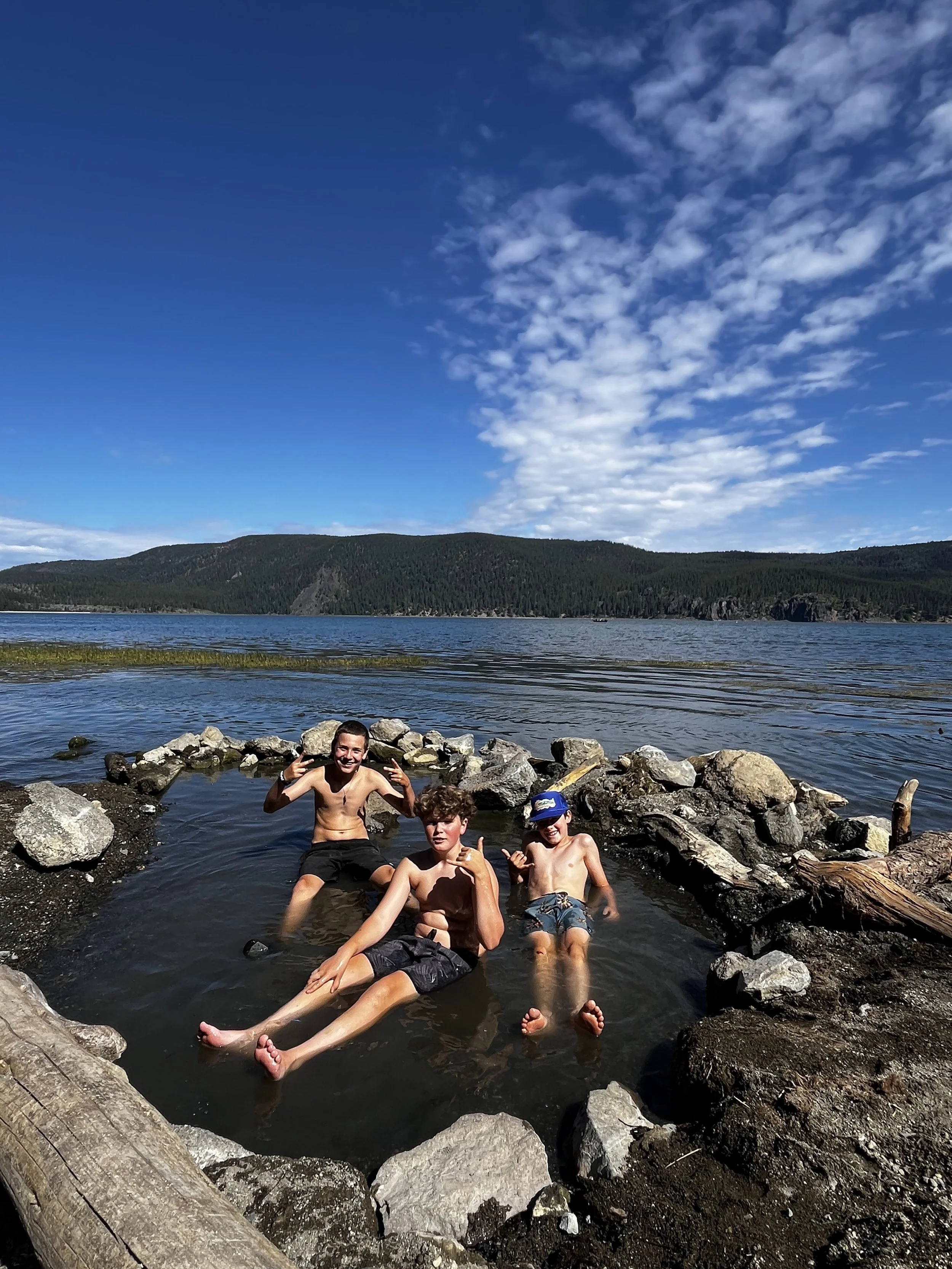 Three boys sitting in a small rocky hot spring pool by the lake with mountains and blue sky in the background.