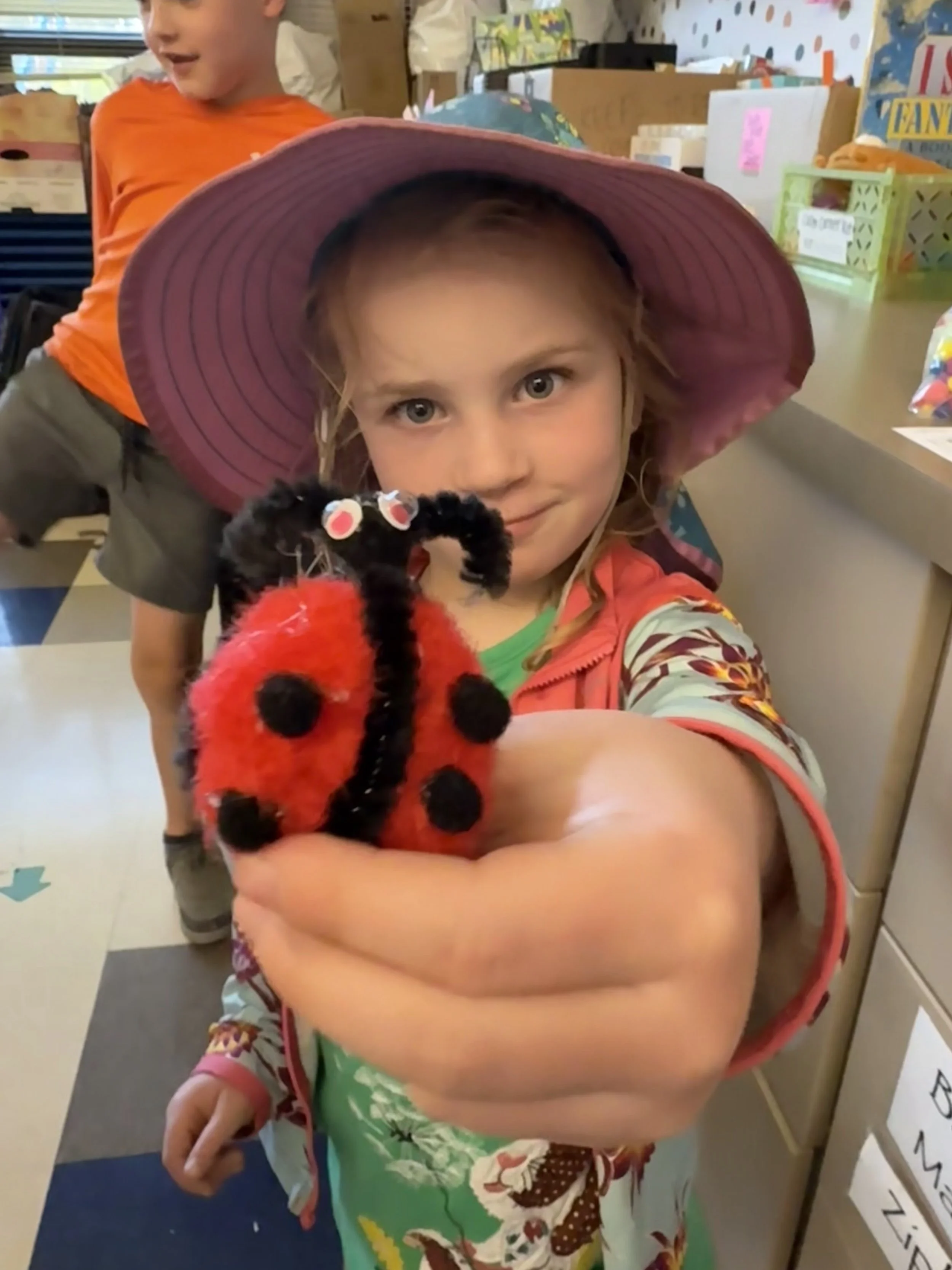 A young girl in a pink sunhat is holding a plush ladybug toy, looking at the camera with a smile, while a boy in an orange shirt stands in the background.