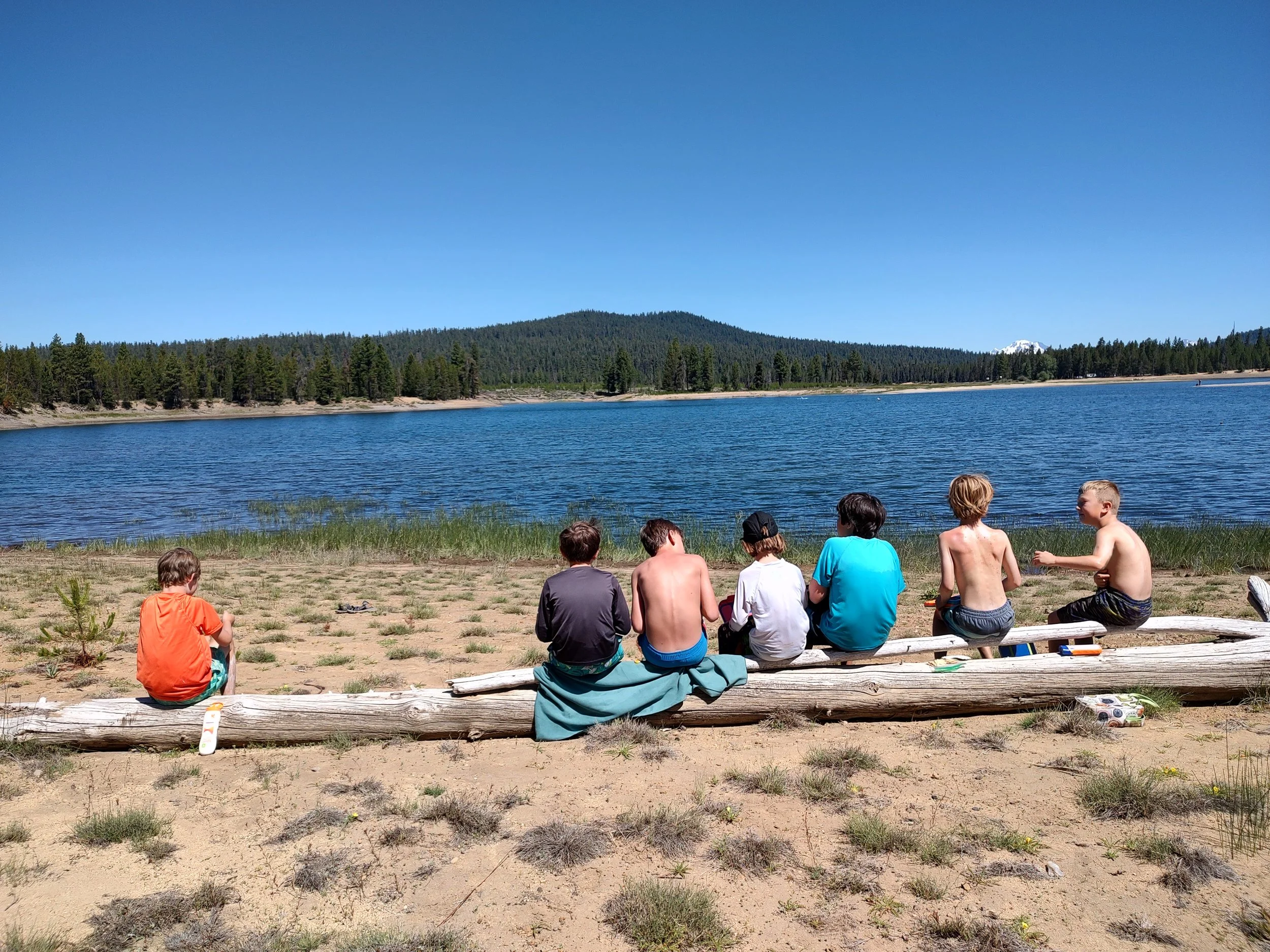 Group of children sitting on a log at the edge of a lake, with pine trees and mountains in the background under a clear blue sky.