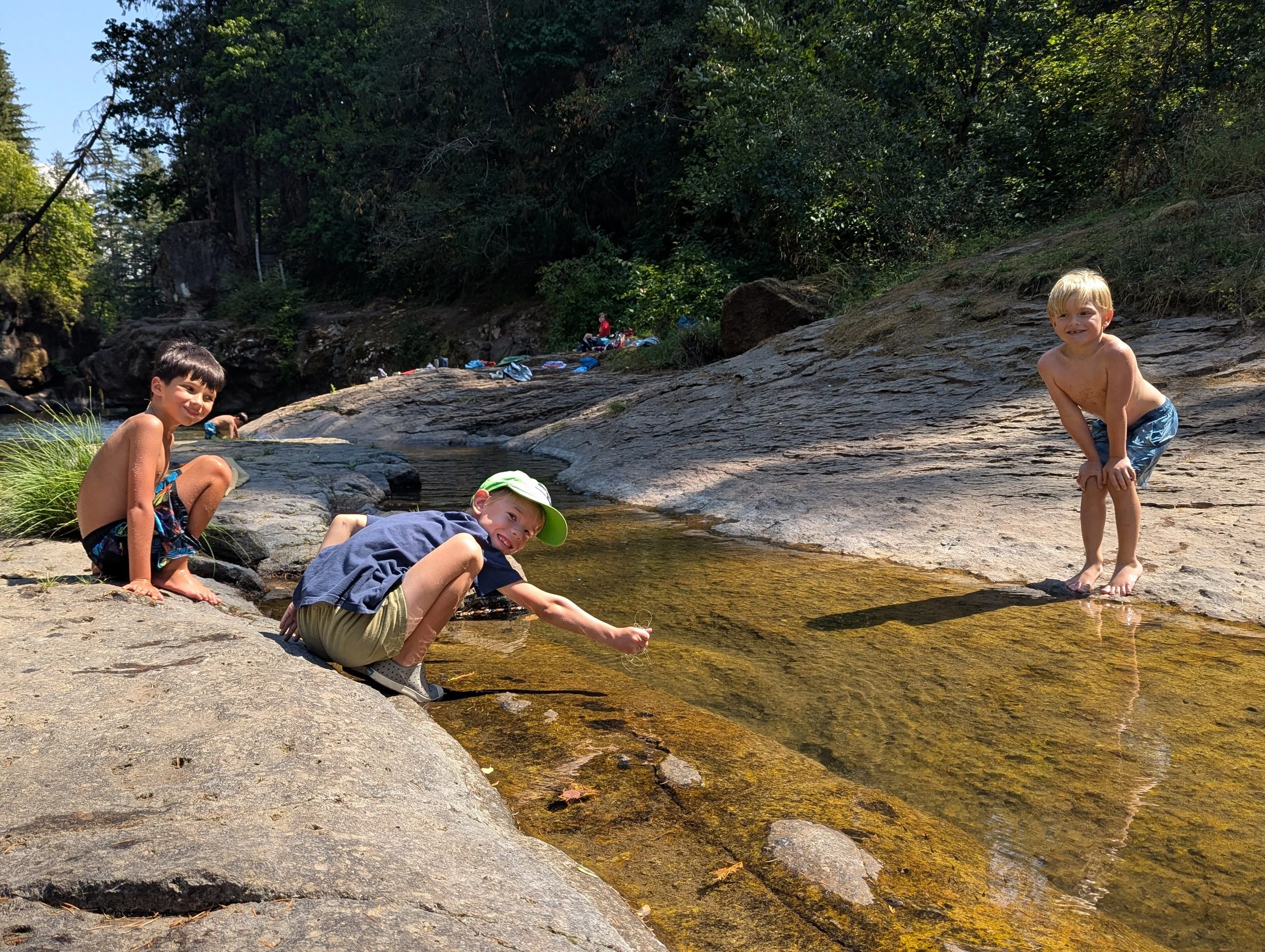 Three children playing and sitting near a small stream in a wooded outdoor setting, with other people in the background.
