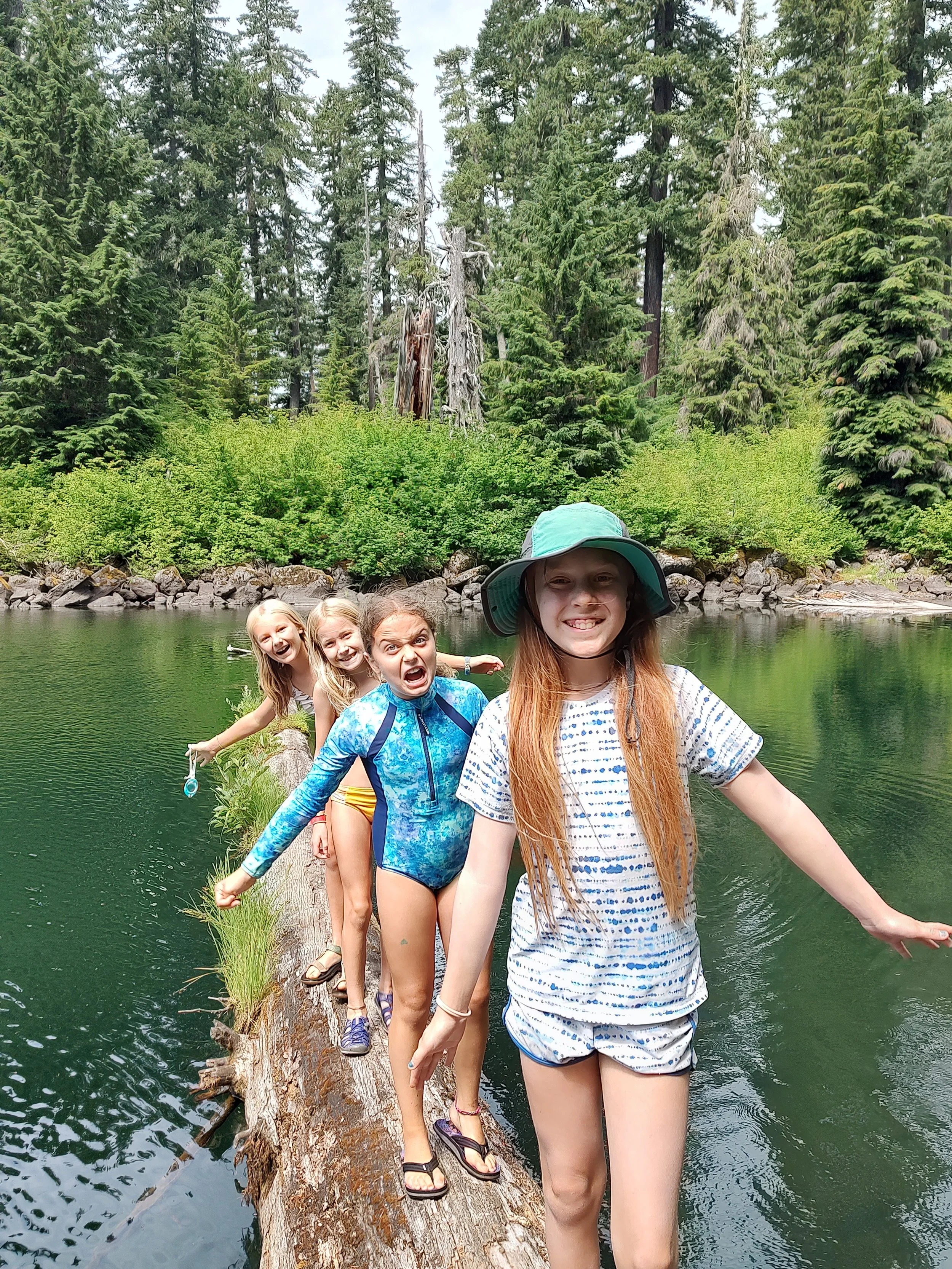 Four children walking on a fallen log across a lake in a forest, with tall pine trees and green foliage in the background.