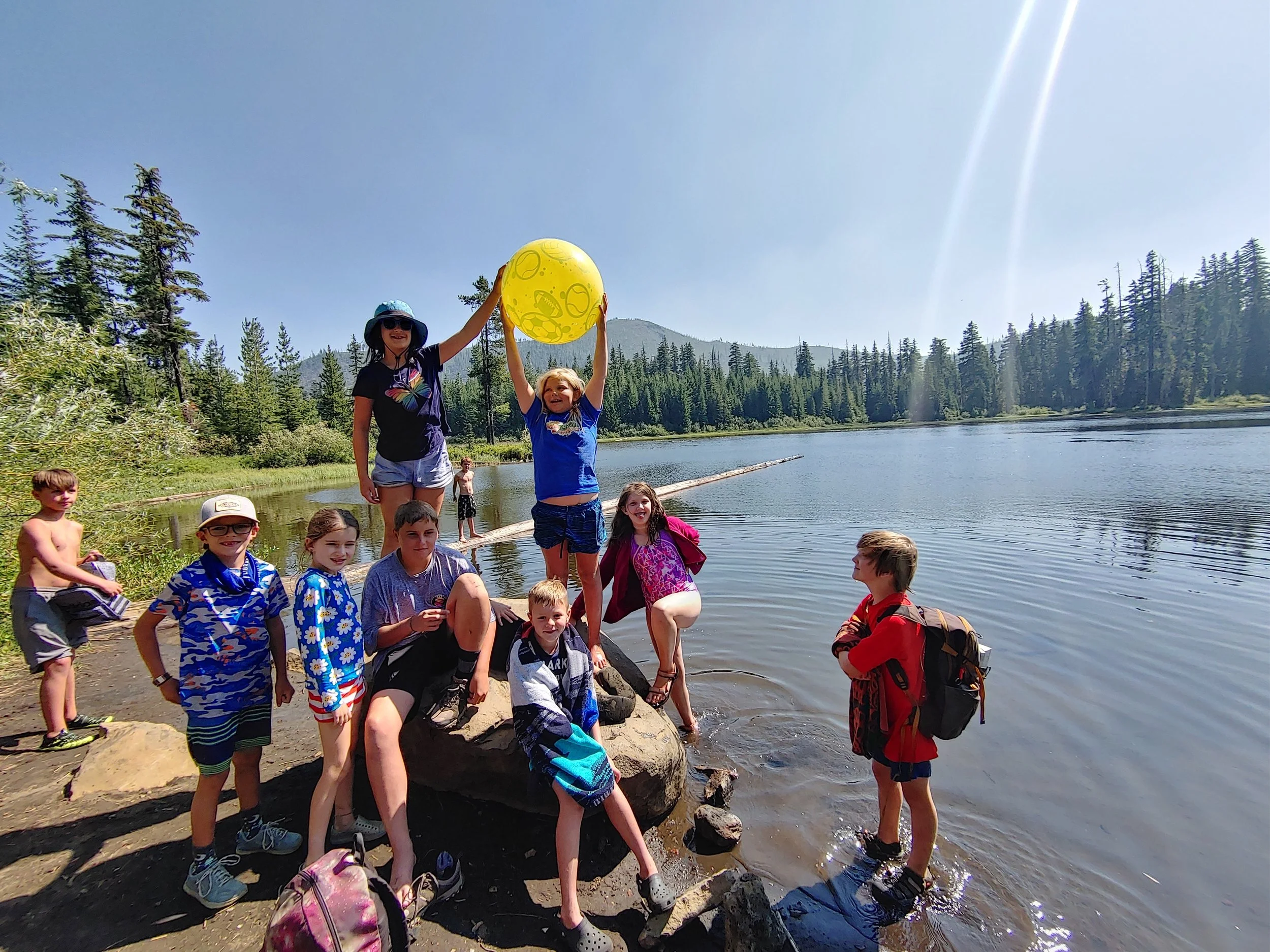 Group of children and a woman by a lakeshore on a sunny day, some children holding a yellow beach ball, with forested hills in the background.