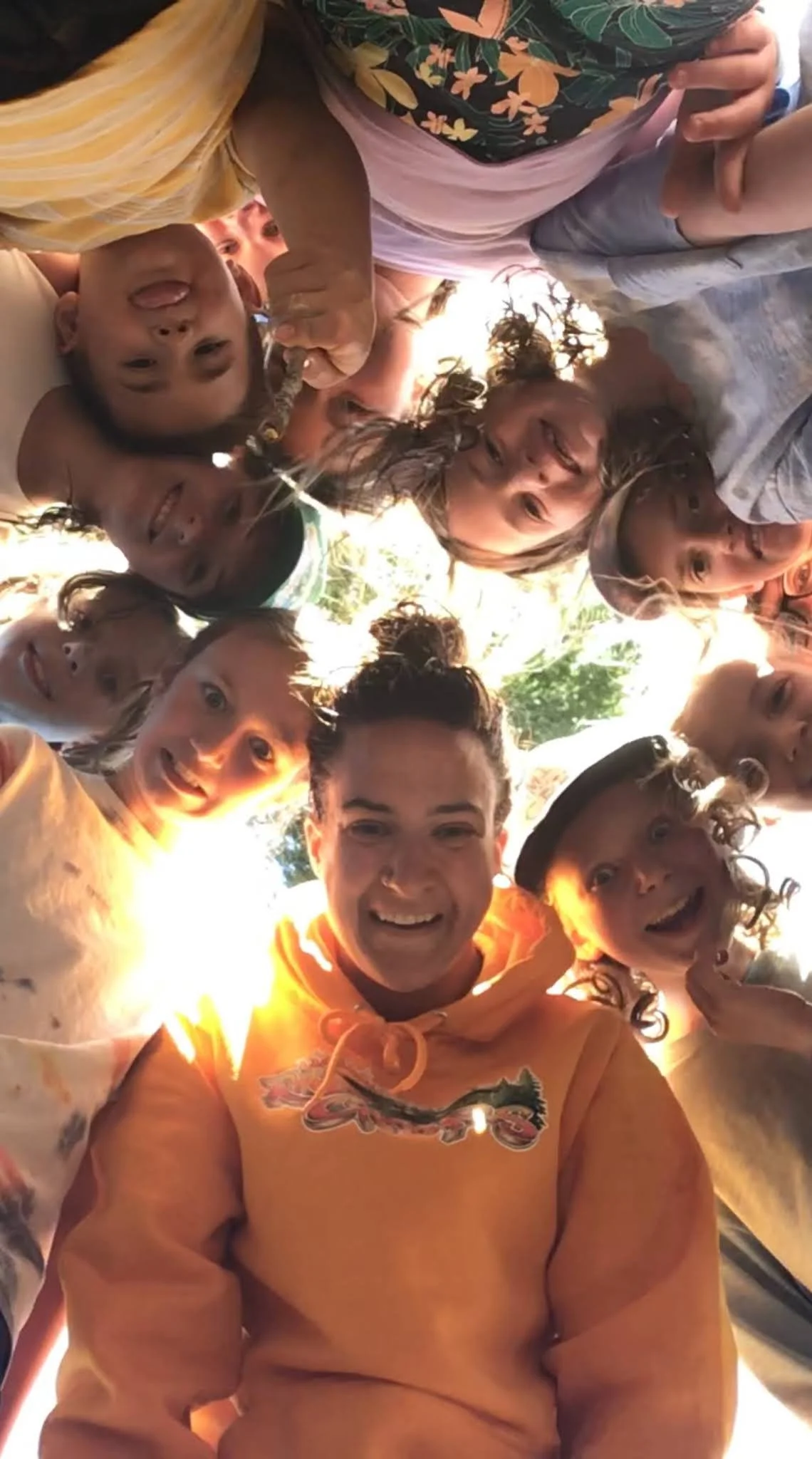 Group of children and young adults smiling and looking down into the camera, taken from below as they gather in a circle outdoors in bright sunlight.