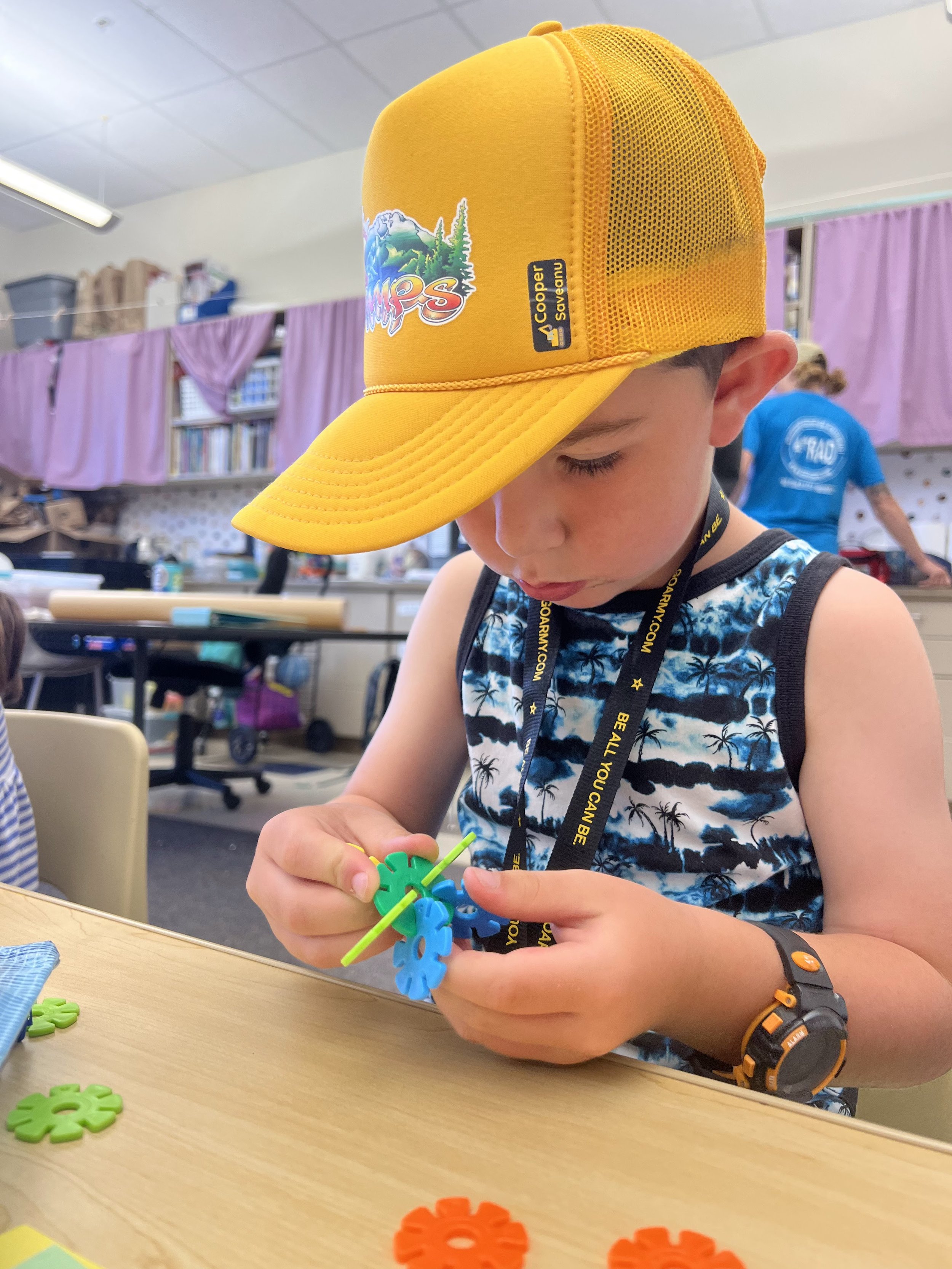A young boy wearing a yellow cap and a sleeveless shirt with a tropical pattern is playing with colorful interlocking pieces at a table, with craft supplies and another child visible in the background.