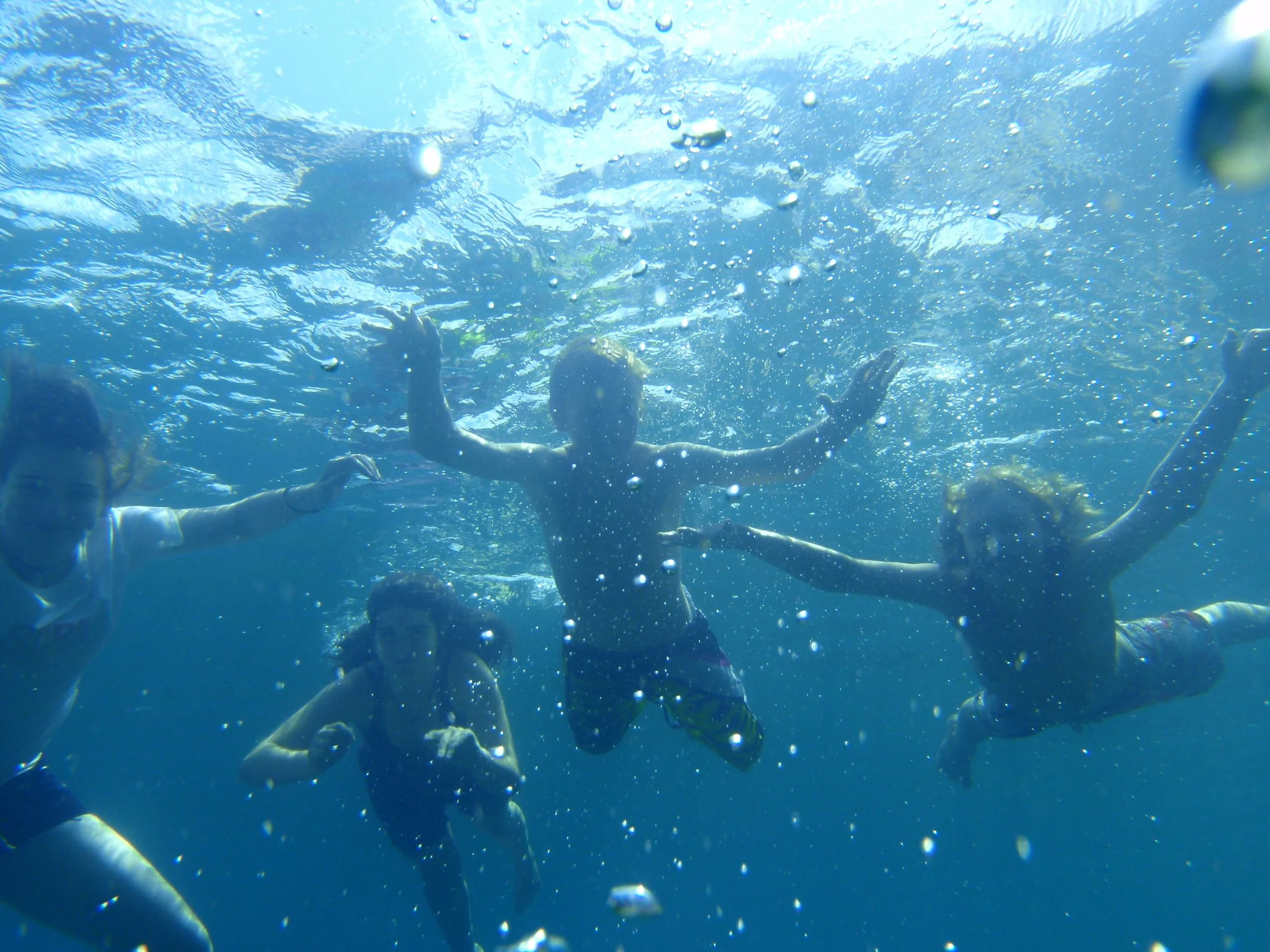 Four children swimming underwater, holding hands and playing in the water, with bubbles around them.