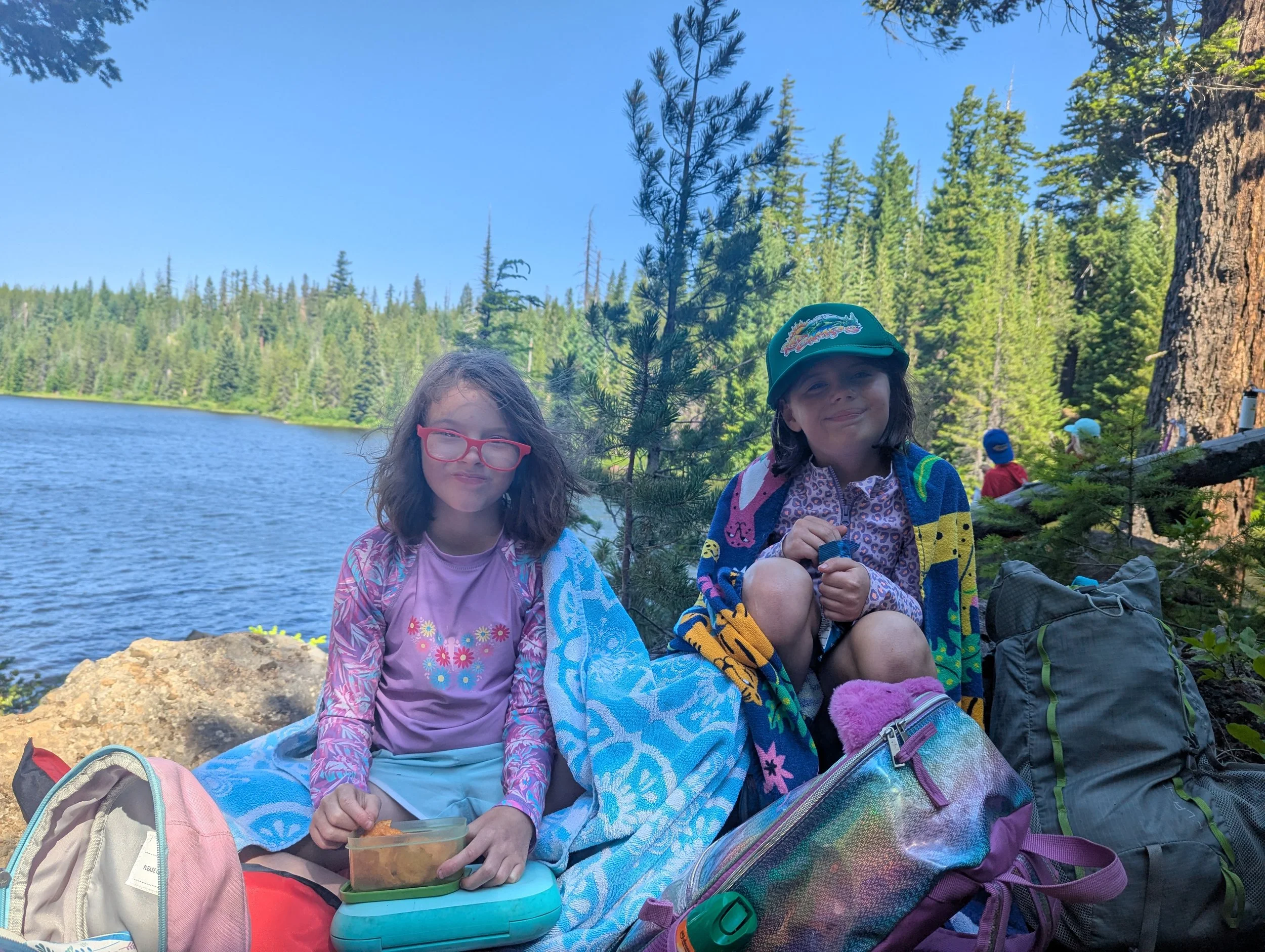 Two young girls sitting on rocks by a lake with a forest background. They are wrapped in colorful towels, wearing hats and glasses, and have backpacks and a lunch container with food.