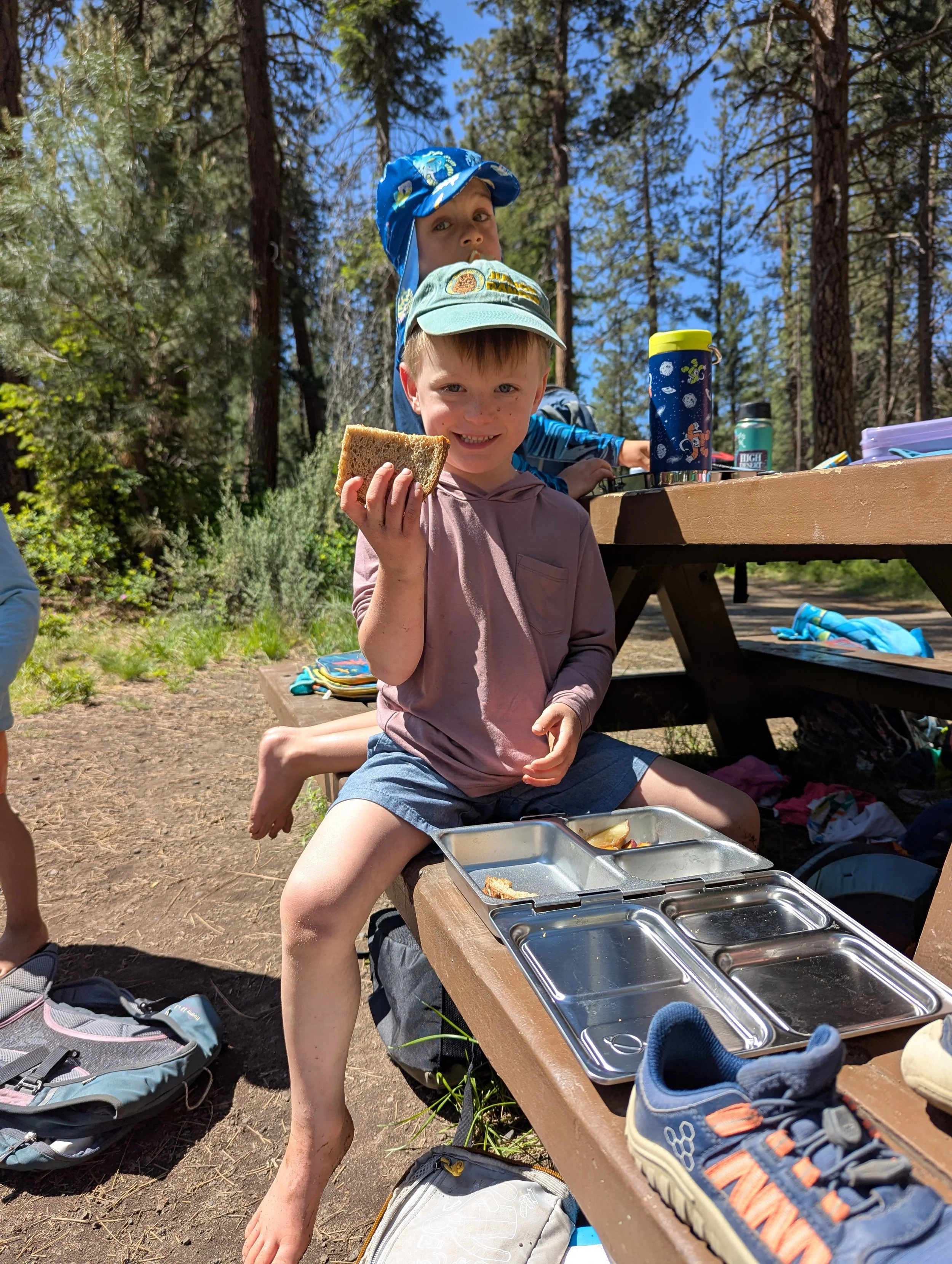 Two young boys enjoying a picnic outdoors in a forest with tall trees. One boy is holding a sandwich and smiling at the camera, sitting on a picnic bench, while the other boy is behind him, slightly higher, wearing a blue hat and looking towards the 