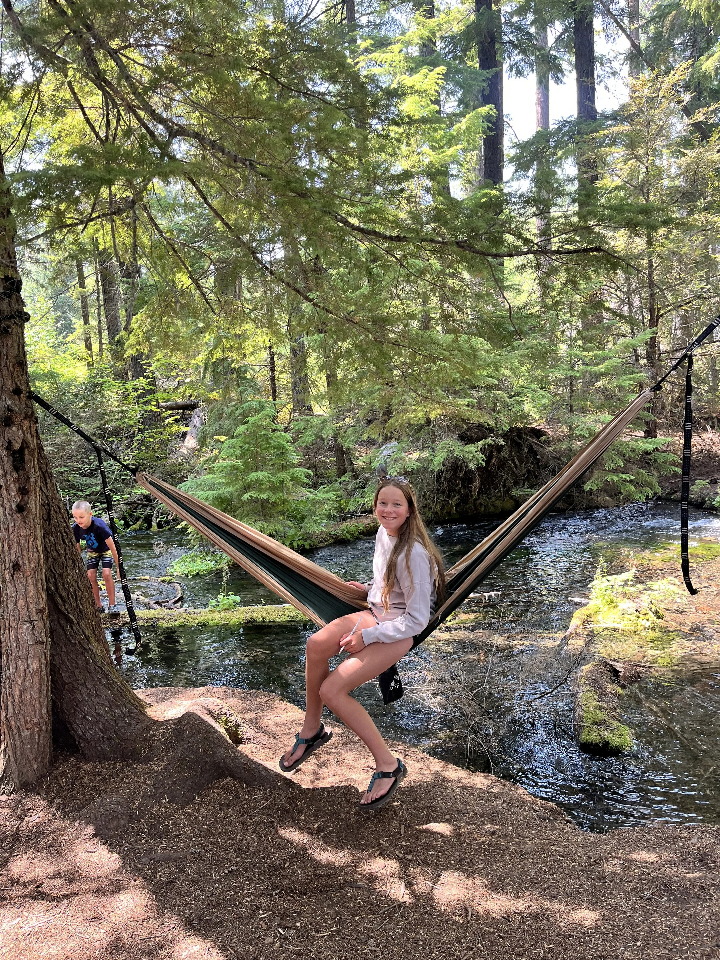 A girl sitting on a hammock tied between two trees in a forest near a creek, with a boy playing in the water nearby.
