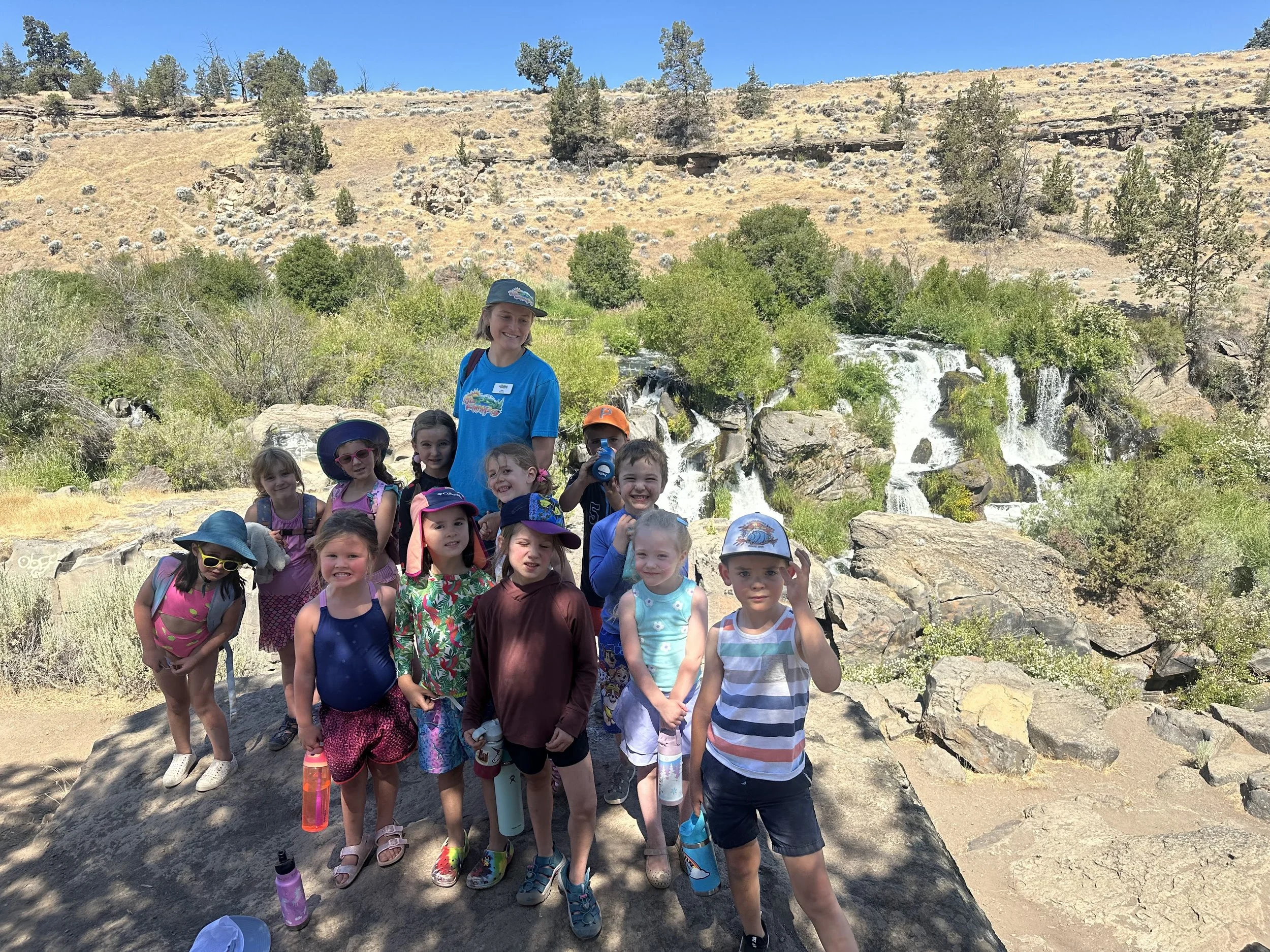 A group of young children and a female adult pose for a photo outdoors near a waterfall, with hills and trees in the background on a sunny day.