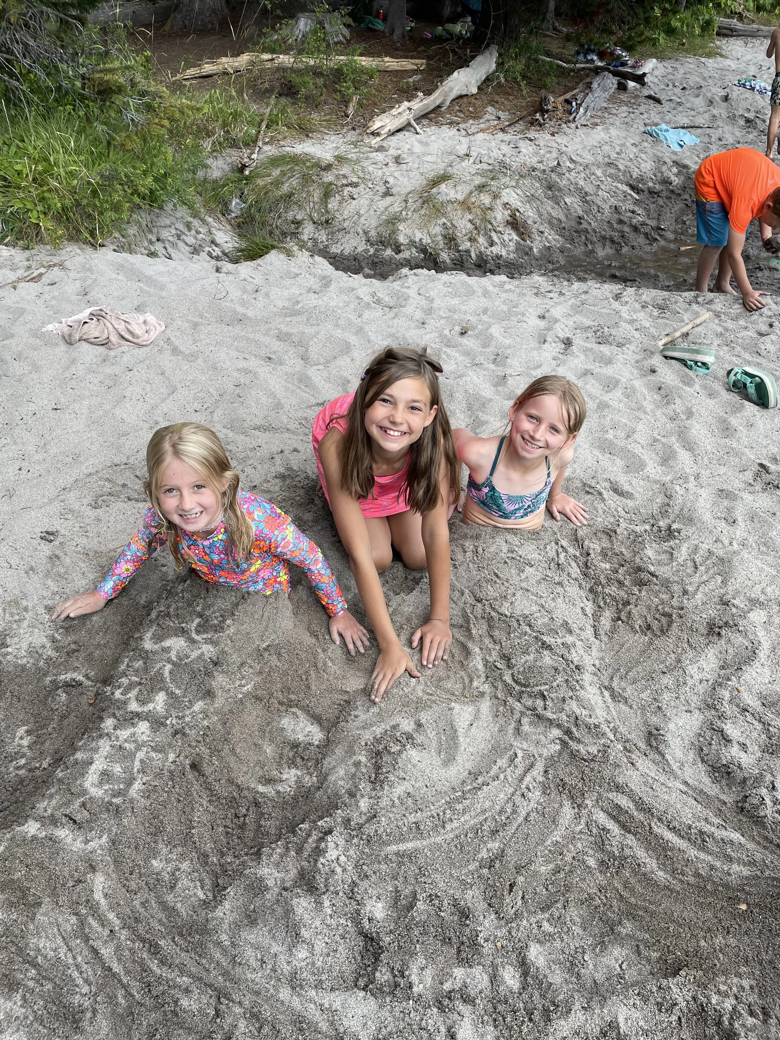 Three young girls sitting in a hole on a sandy beach, smiling at the camera. In the background, a man in an orange shirt is bending down near the shoreline, and there are some trees and scattered items on the beach.