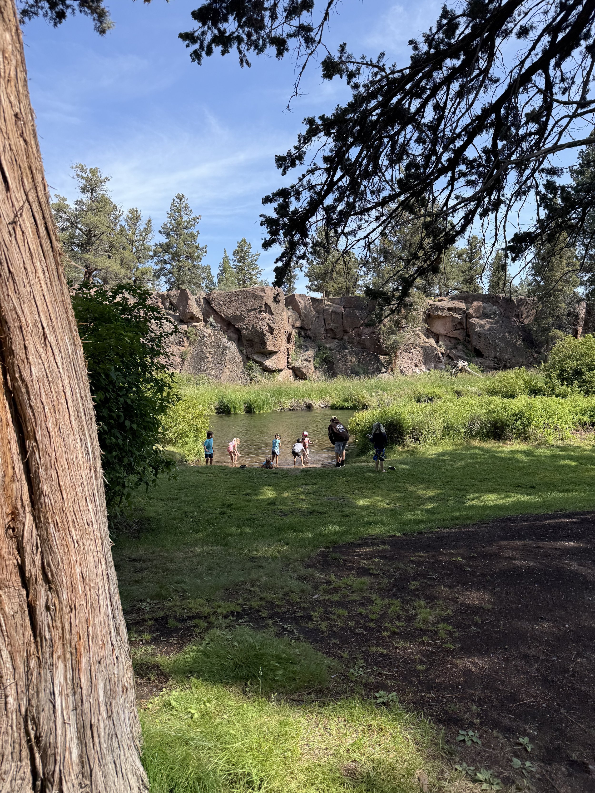 Family and children standing by a river in a forested area, with large rocks and trees in the background.
