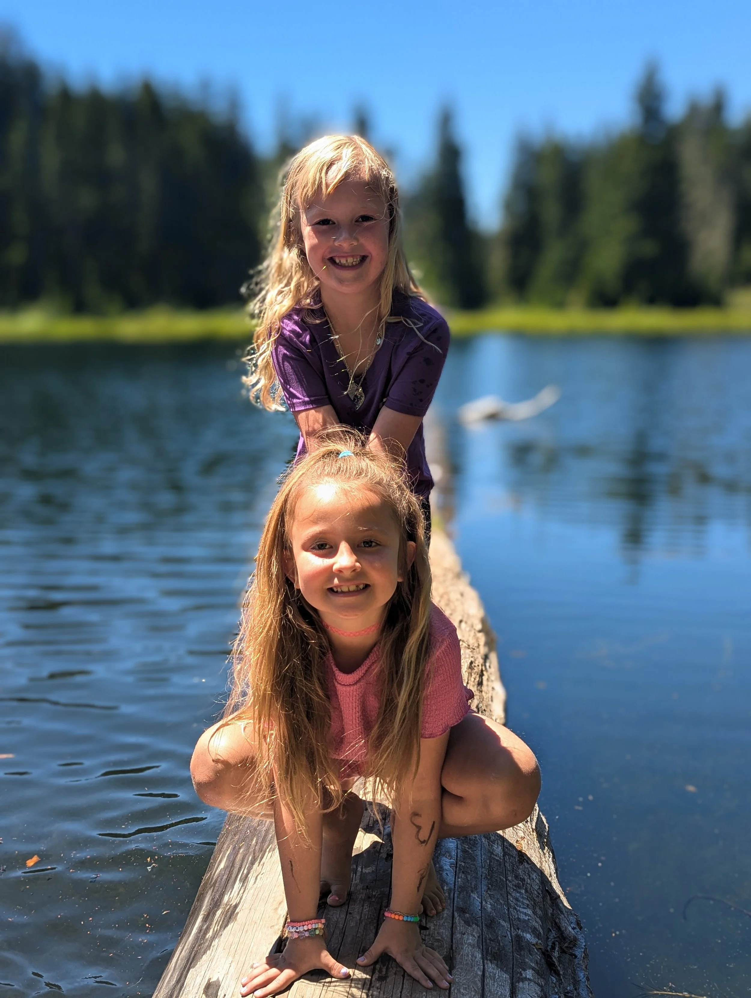 Two young girls with long blonde hair squatting and leaning on a wooden log at a lakeside, surrounded by trees with a clear blue sky overhead.