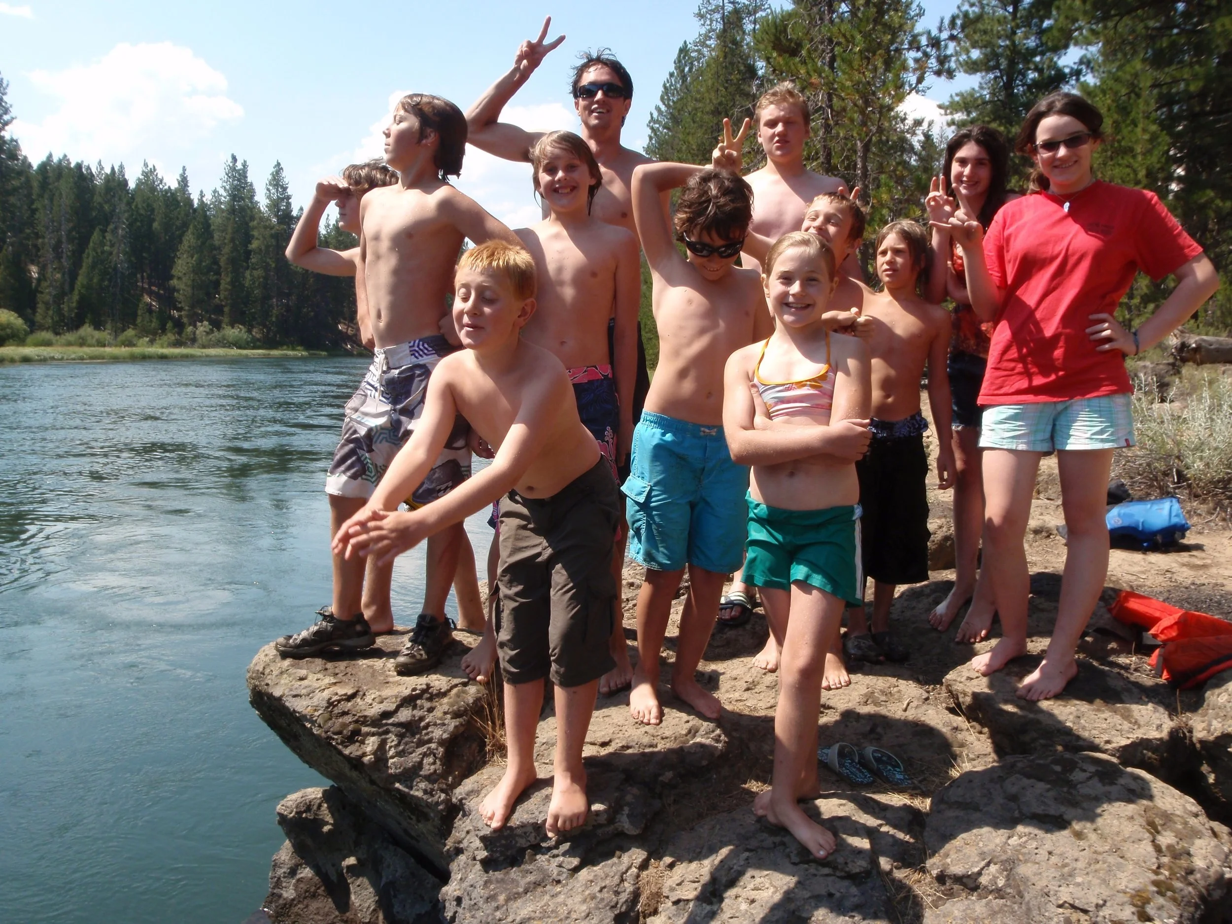 A group of children and teenagers along with an adult posing on rocks beside a lake in a forested area, some in swimwear and making peace signs.