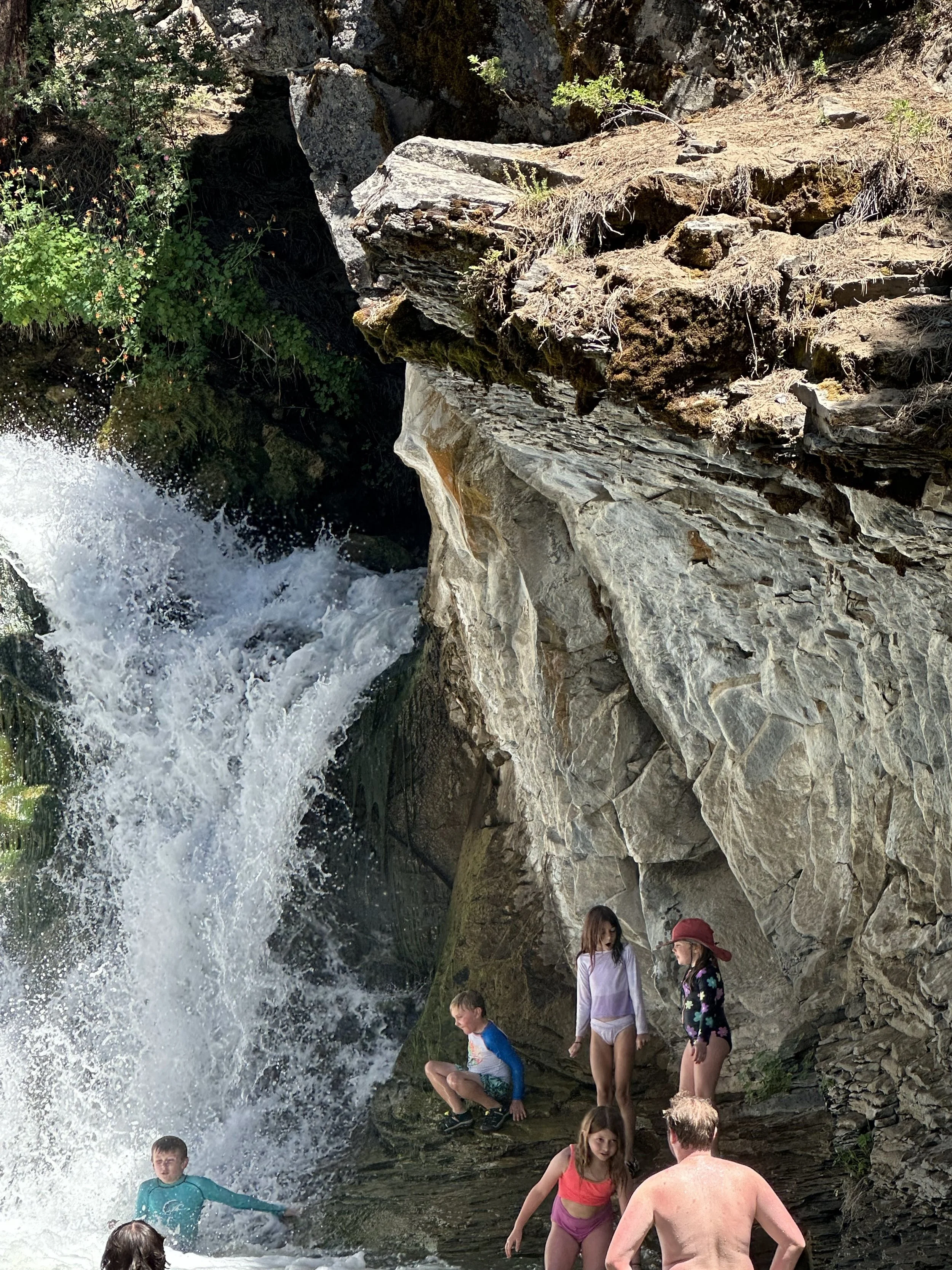 Children swimming and playing near a waterfall in a rocky outdoor setting.