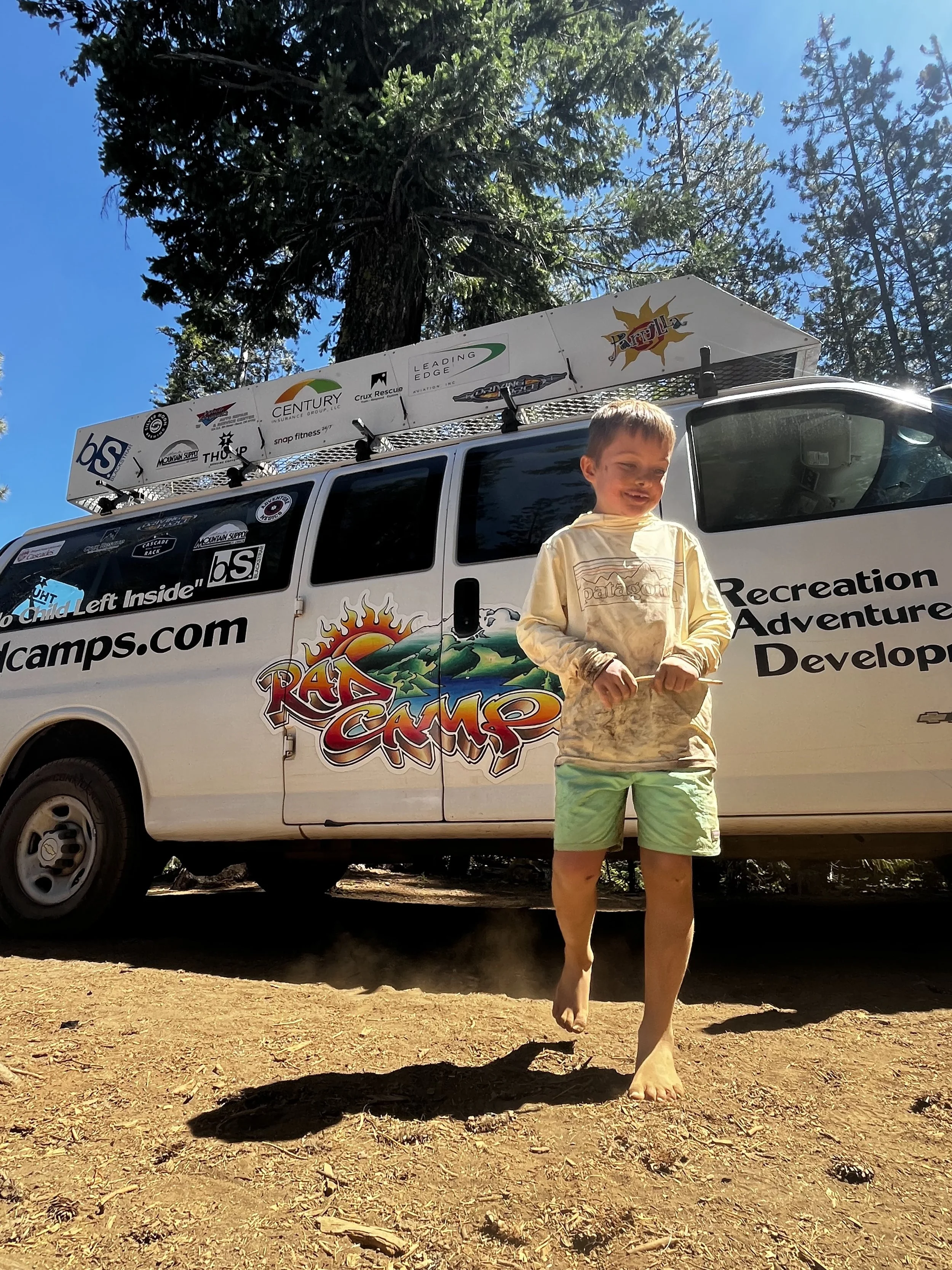 A young boy running barefoot in a dirt area in front of a white recreation van with colorful camping logo and tree graphics, surrounded by tall pine trees with a clear blue sky overhead.