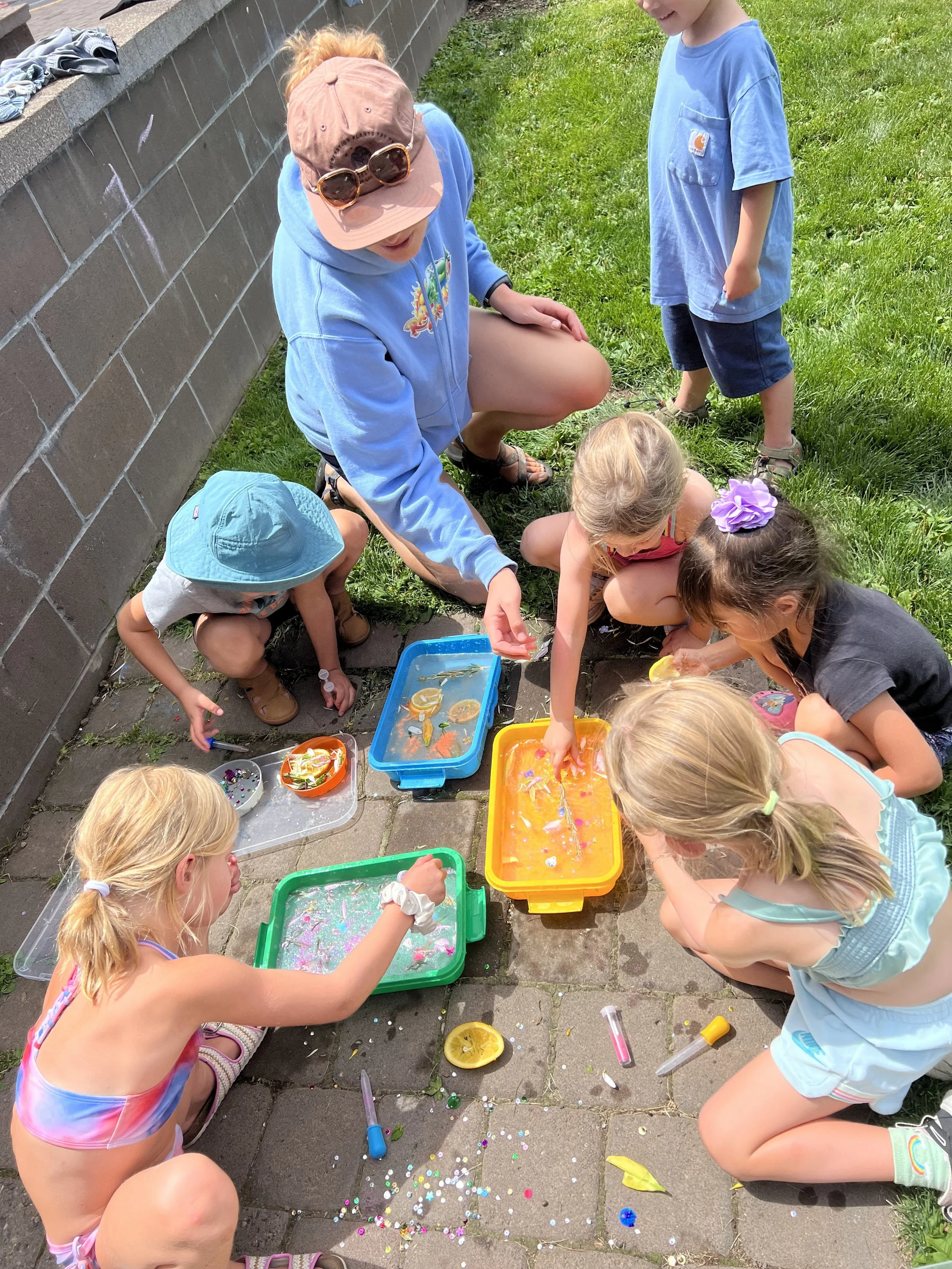 Children playing with water and floating objects outdoors on a sunny day, sitting and kneeling on the ground near a brick wall and grass.