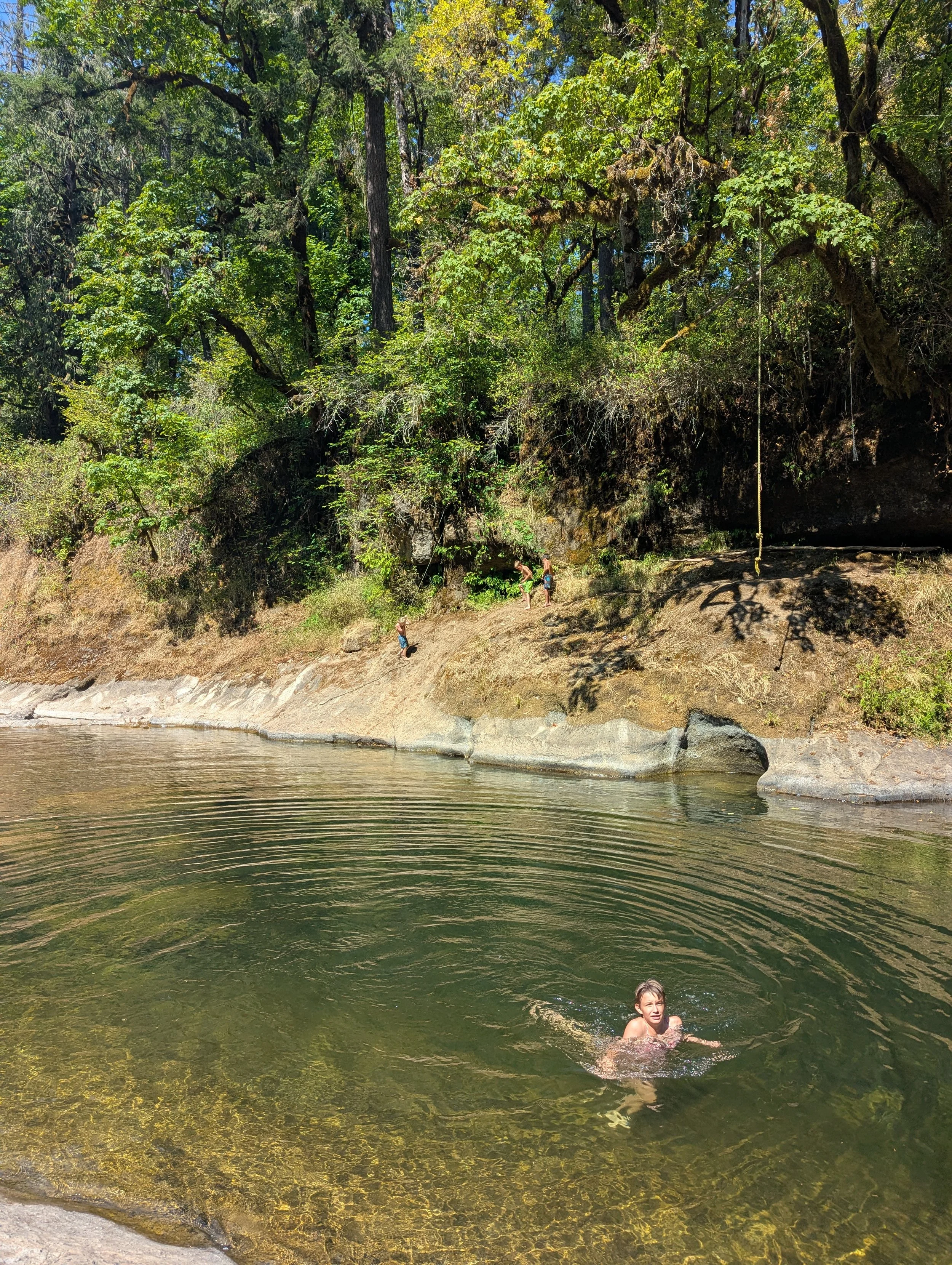 A person swimming in a natural river with a rocky bank, surrounded by lush green trees and cliffs, with a group of people near the shore.