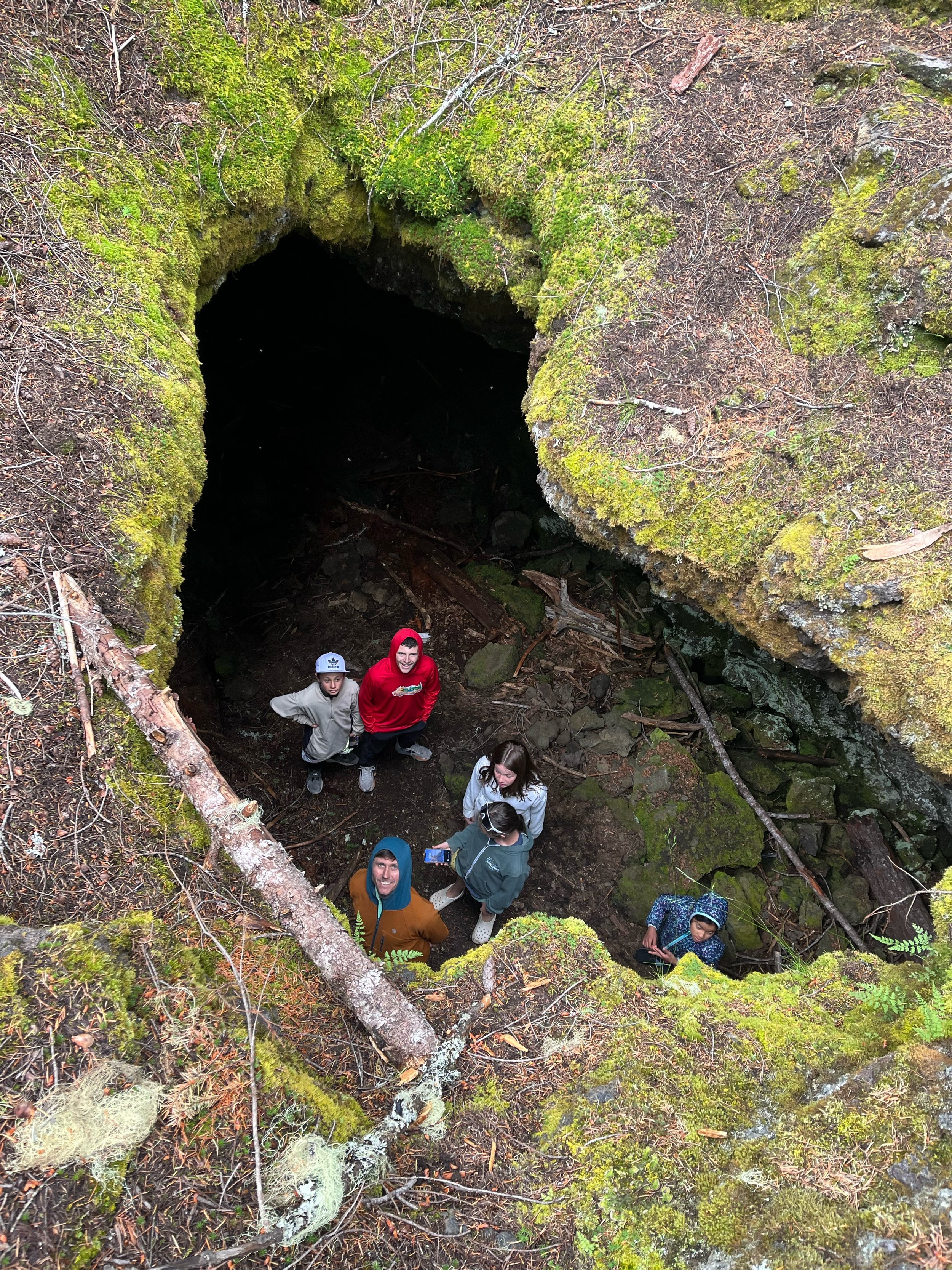 A group of people standing at the edge of a large moss-covered cave opening in a forest.