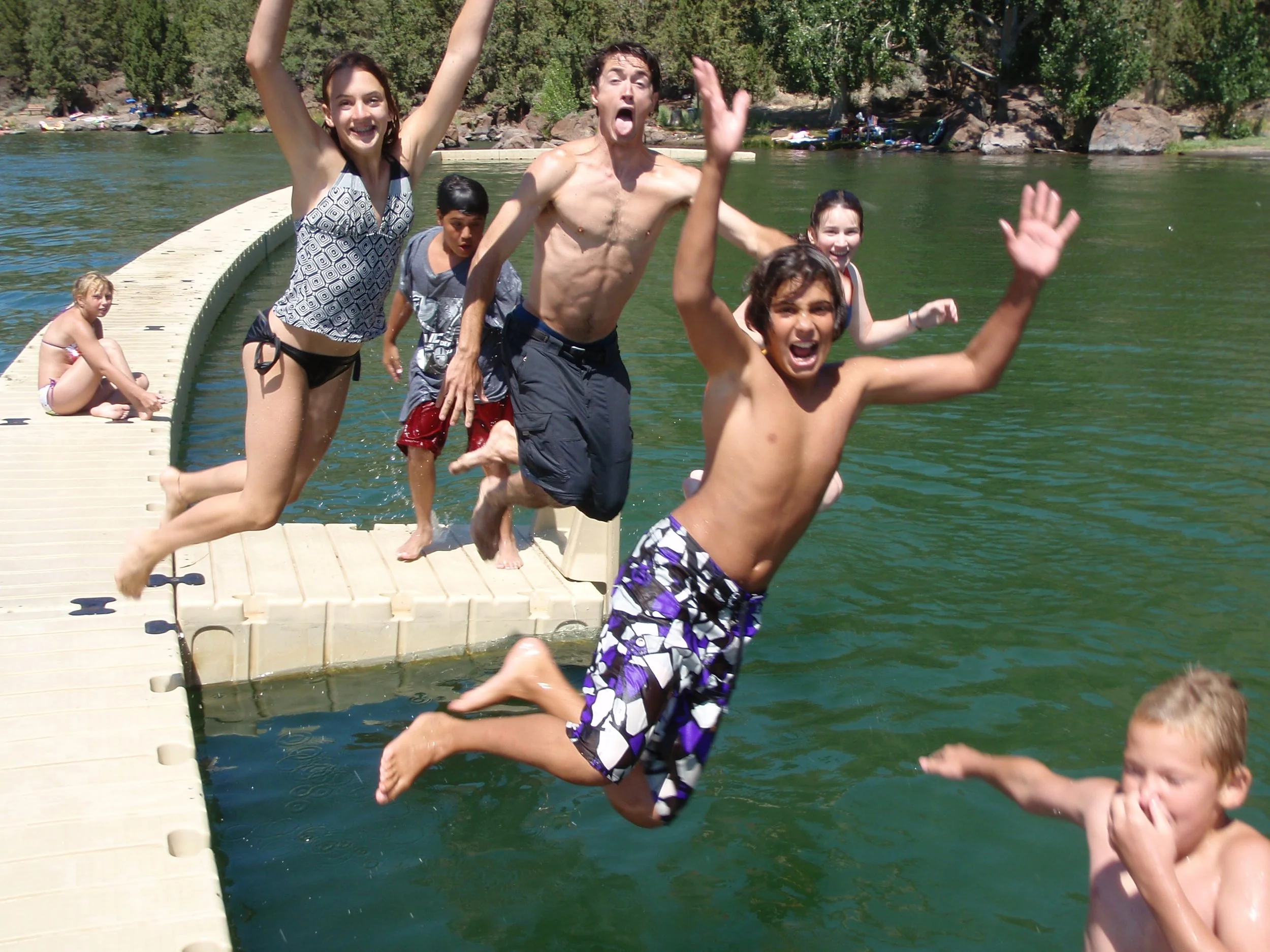Group of children and teenagers enjoying a day at the lake, jumping into the water from a dock on a sunny day.