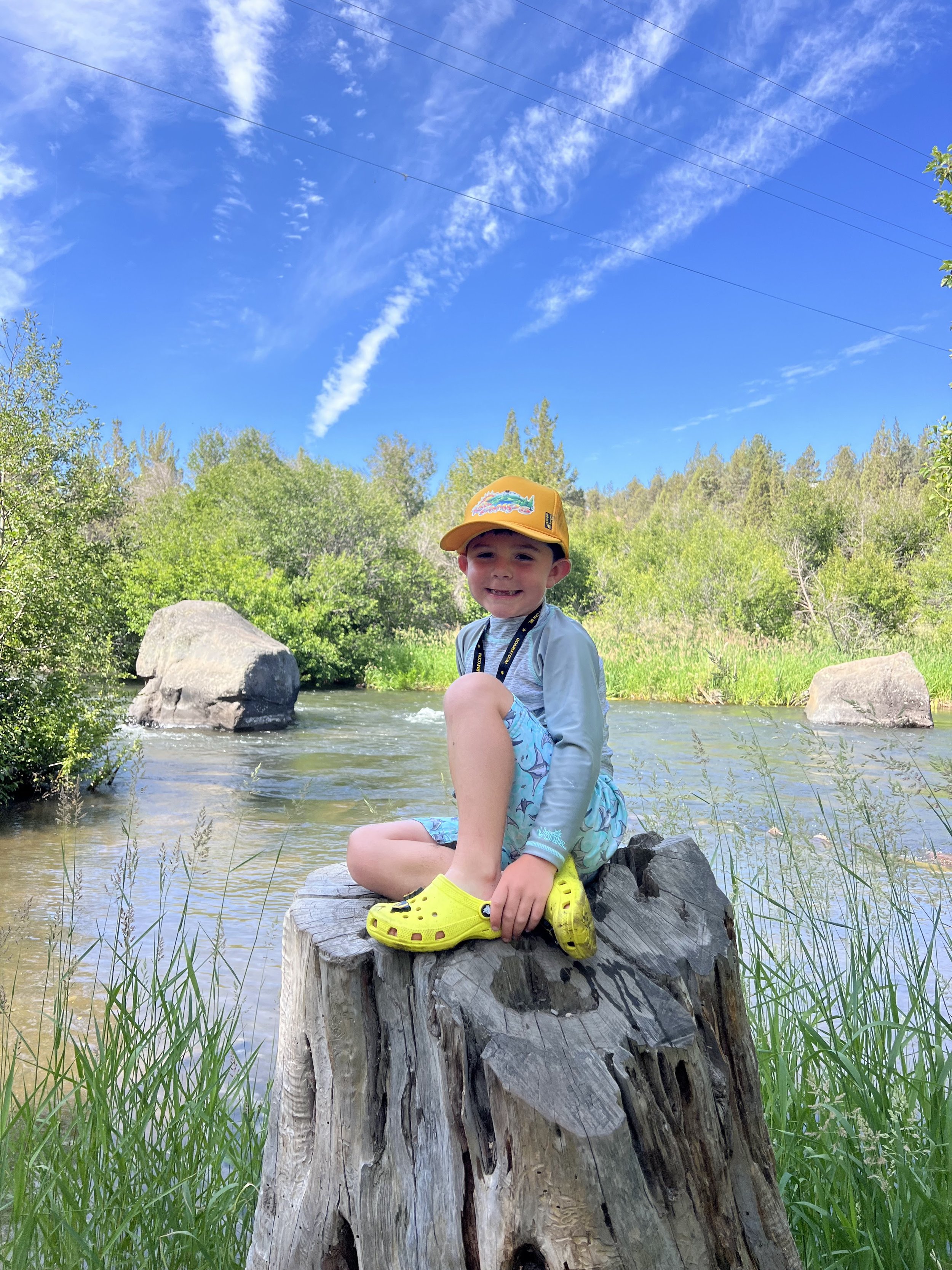 A smiling young boy sitting on a tree stump in a creek, wearing a yellow cap, a light blue jacket, colorful shorts, and bright yellow Crocs, with green trees and a blue sky with clouds in the background.