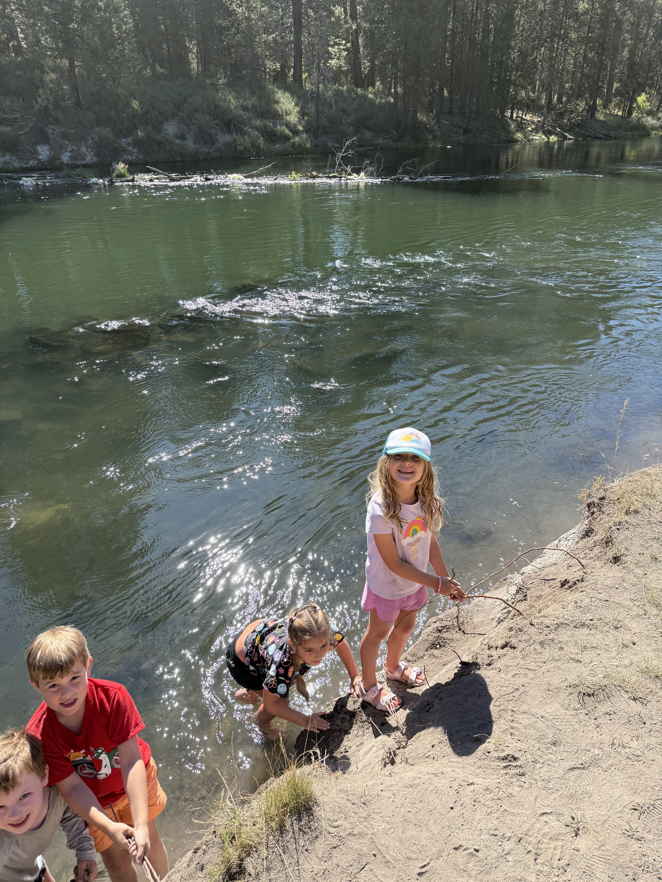 Four children, three boys and one girl, standing on the sandy shore of a river, with some children climbing out of the water, the girl is smiling at the camera, wearing a rainbow t-shirt and cap, while holding a fishing stick. The river is surrounded
