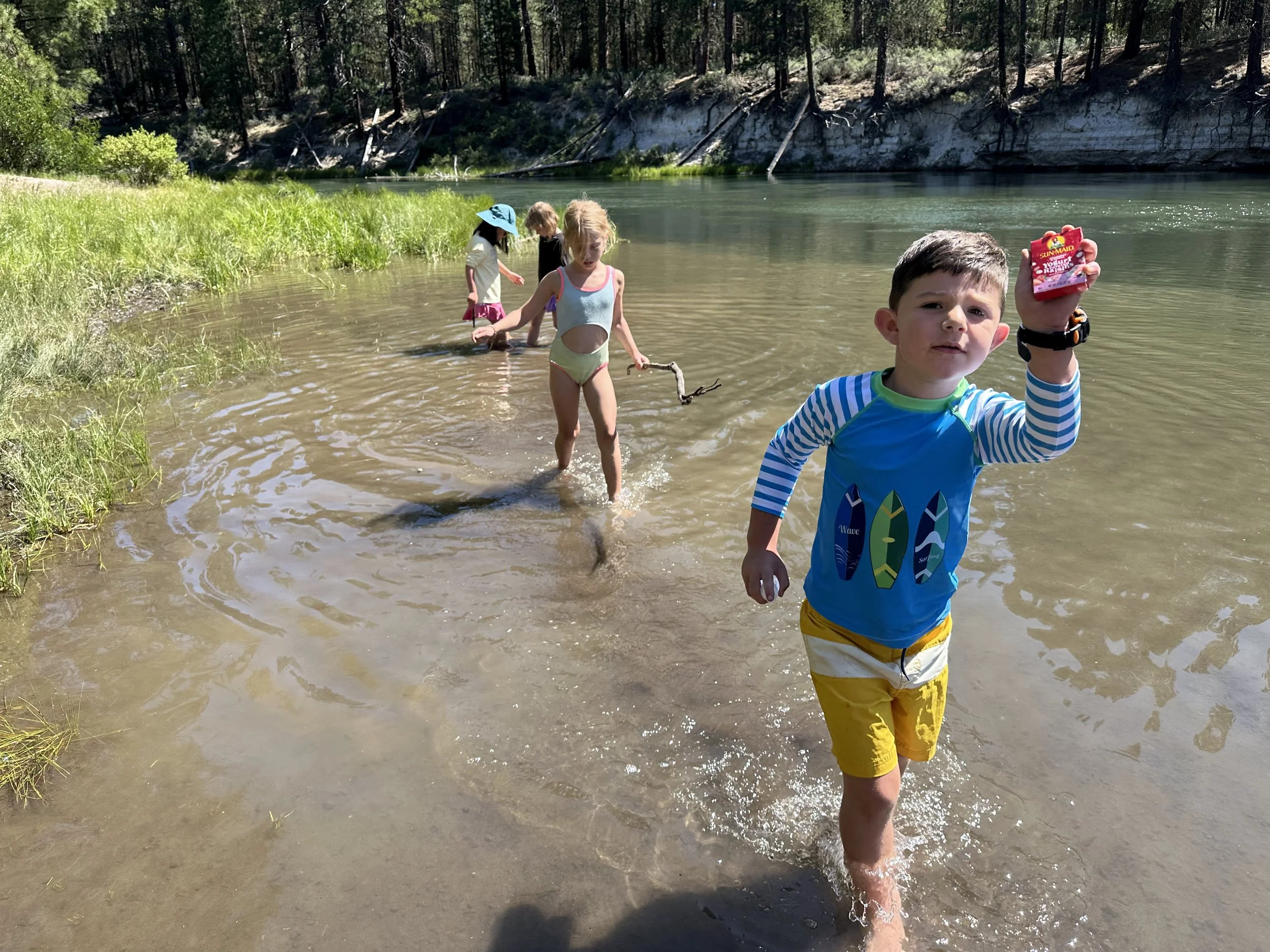 Four children playing in a shallow river near a wooded area during summer, with one boy in the foreground holding a small red toy or object, wearing yellow shorts and a blue shirt with surfboard designs.