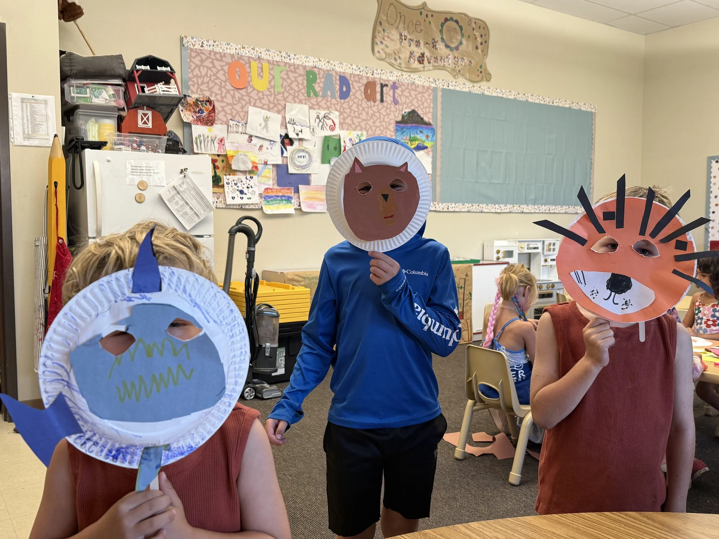 Children in a classroom wearing paper animal masks, including a shark, a cat, and a fox, during an art activity.