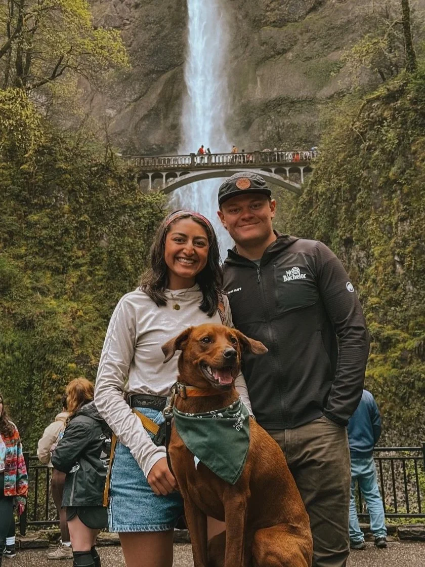 A happy couple with a brown dog in front of a waterfall at a scenic overlook, with other visitors in the background.
