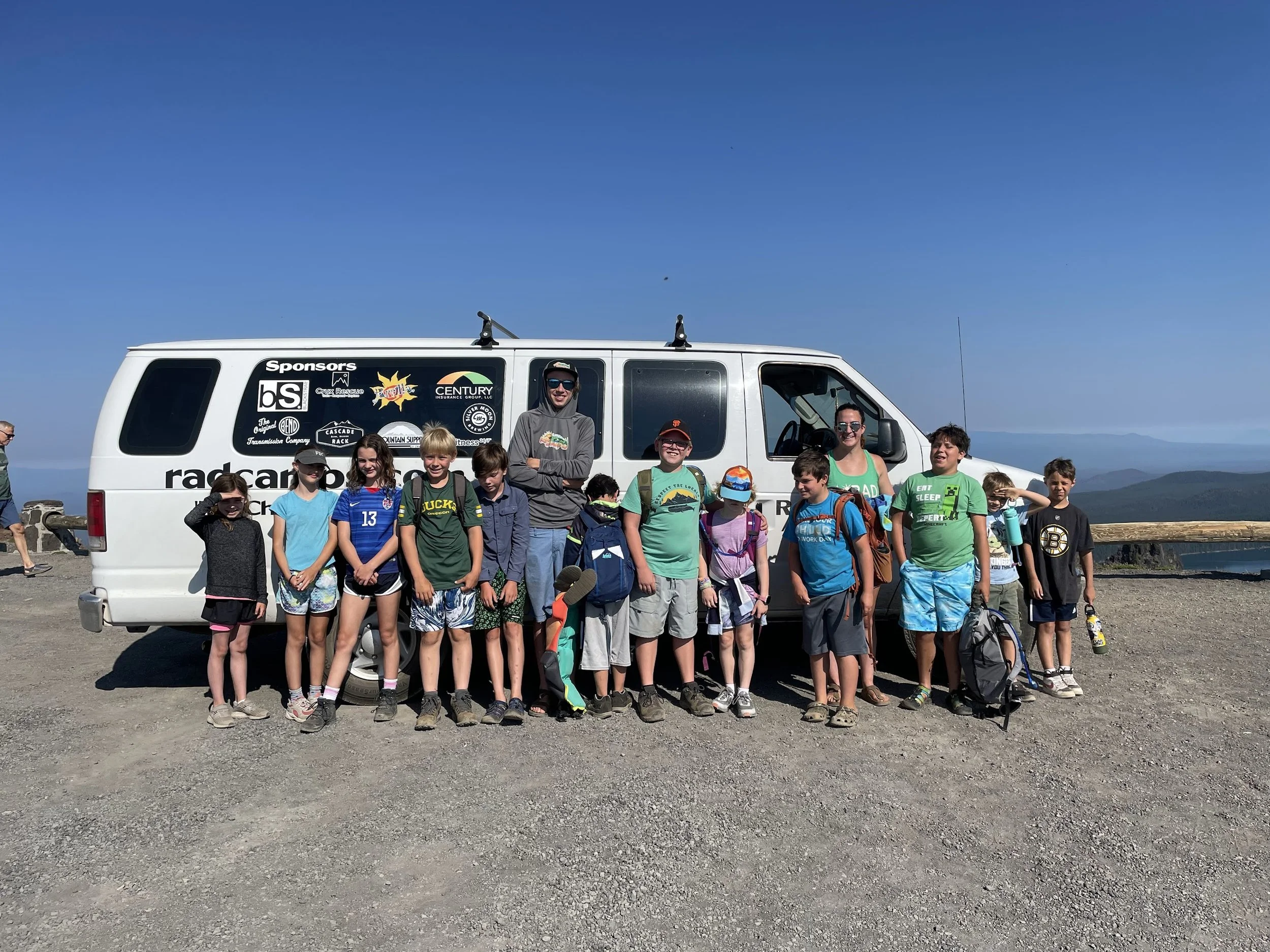 Group of children and two adults standing in front of a white van on a mountain with blue sky and distant mountains in background.
