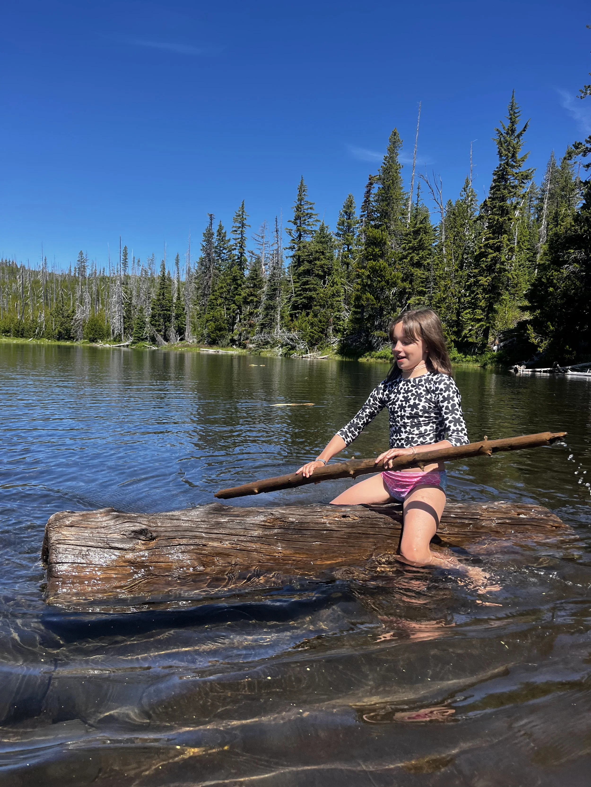 A young girl in a black-and-white patterned long sleeve shirt and pink shorts kneeling on a large log in a lake, holding a long stick, with a forest and blue sky in the background.
