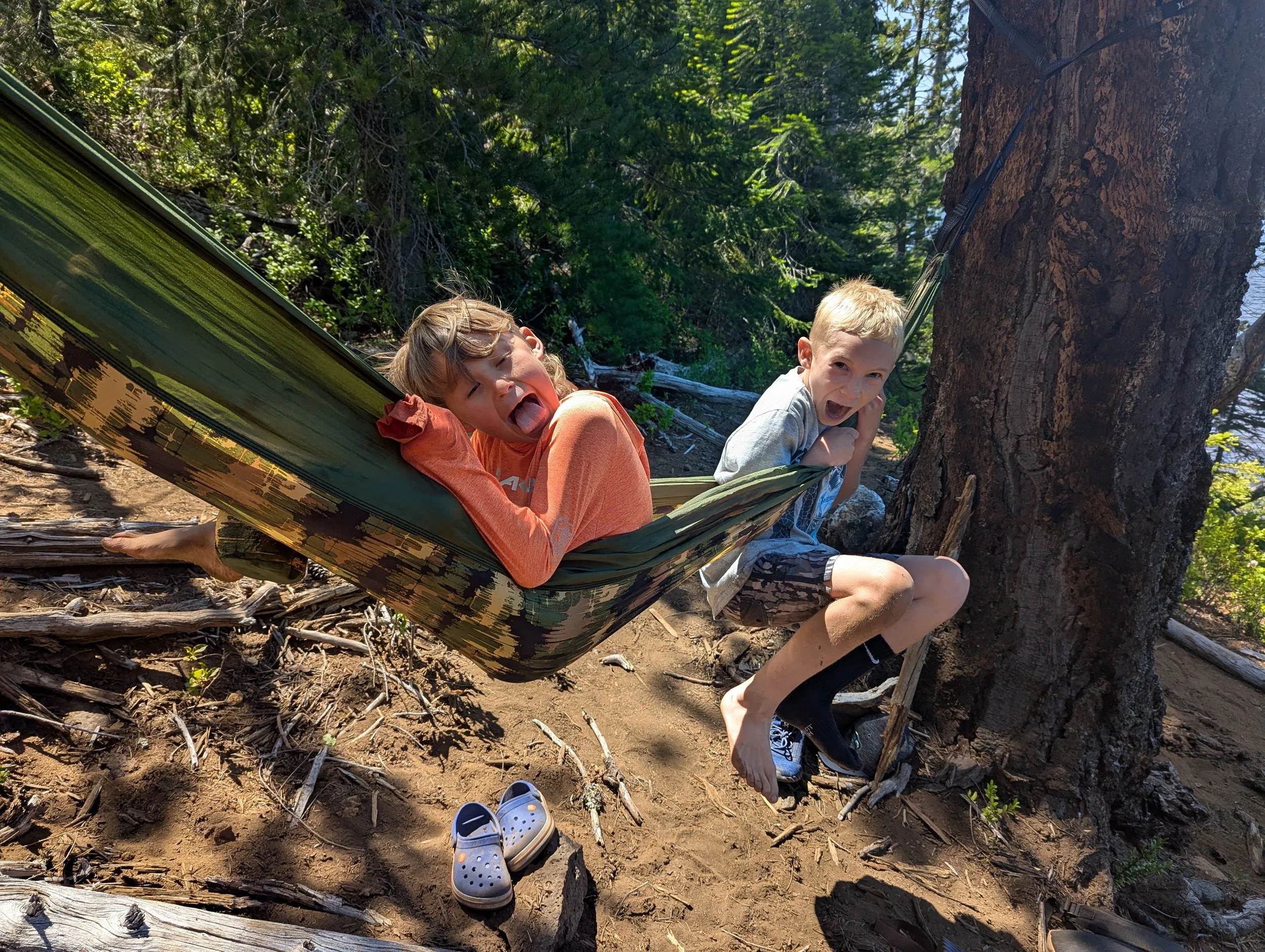 Two children, a girl and a boy, are playing on a hammock tied to a large tree in a wooded area. The girl is lying in the hammock with her tongue out, wearing an orange shirt. The boy is sitting on the ground, smiling and looking at the camera, wearing a gray shirt and black shorts. There are some blue Crocs on the ground near them.