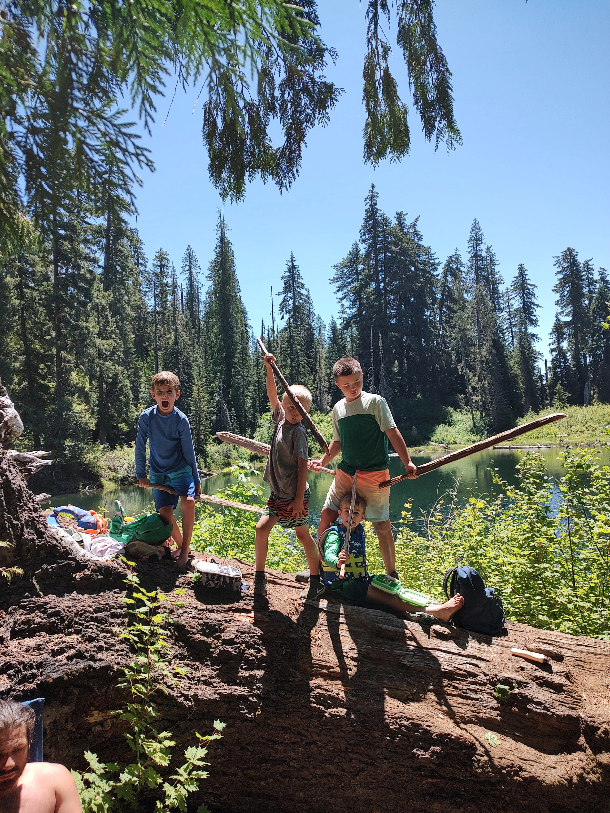 Five children playing on a fallen log by a river in a forest, with tall pine trees and a clear blue sky in the background.