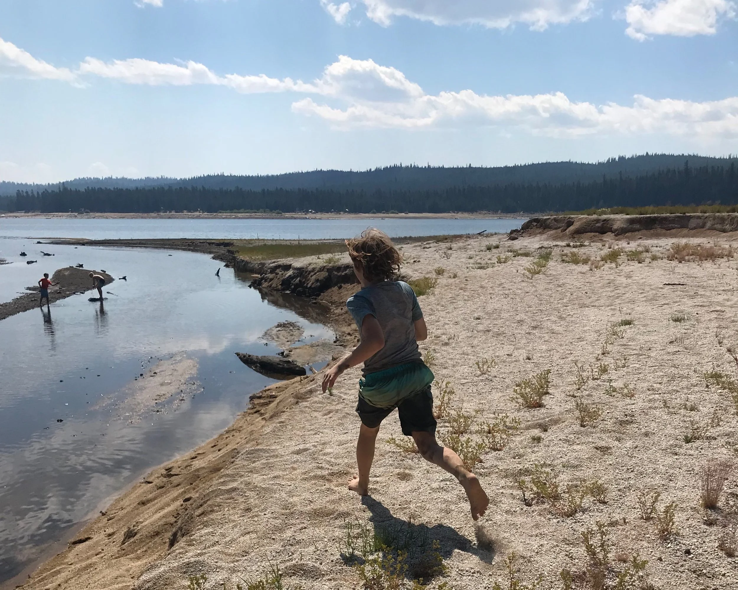 A young boy running on a sandy riverbank near a body of water, with two people wading in the water and a distant forest on the horizon under a partly cloudy sky.